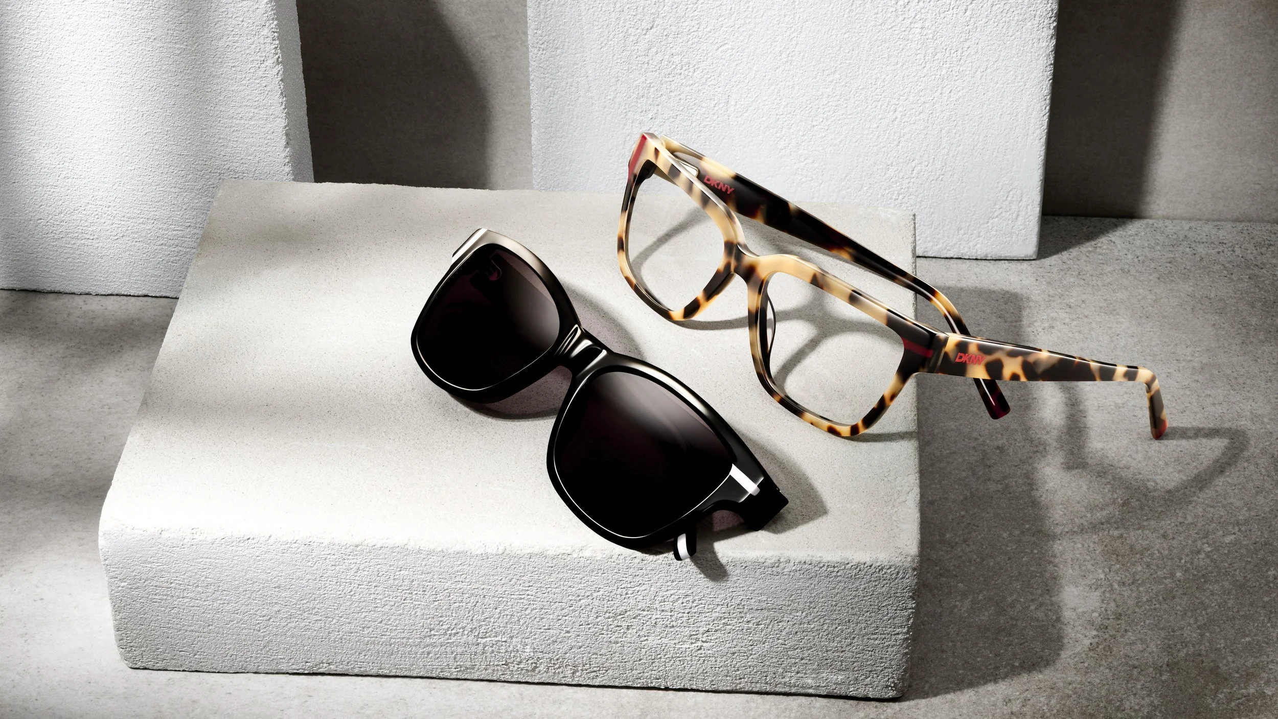 A pair of black sunglasses and a pair of tortoiseshell glasses are displayed on a white, textured block with a concrete background.