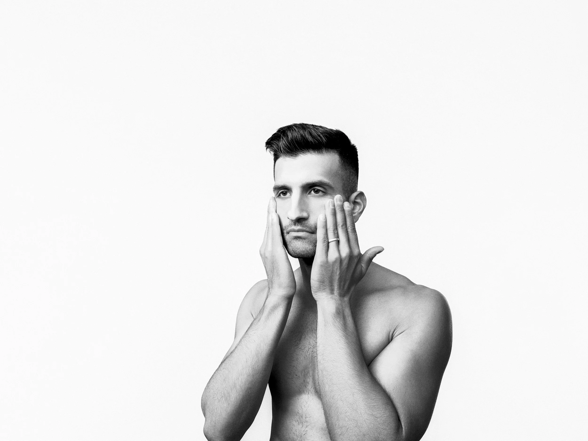 A shirtless man with short dark hair holding his face with both hands against a plain light background.
