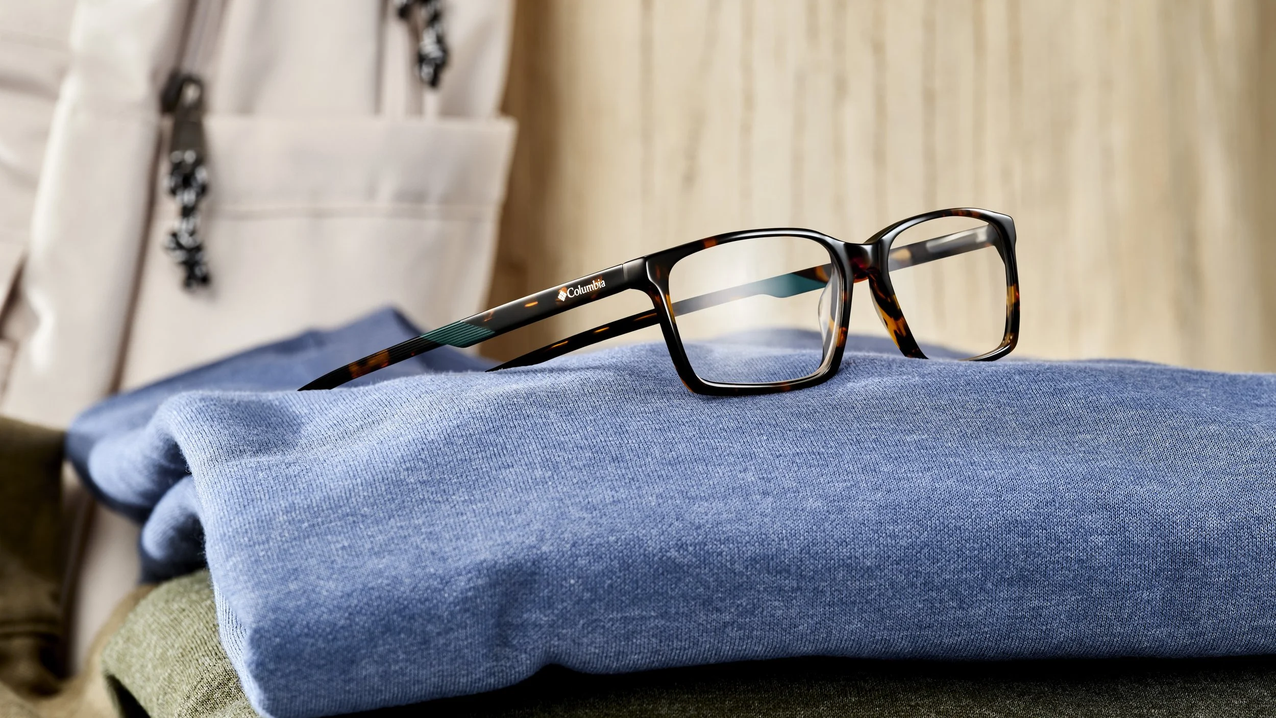 Pair of black and brown tortoiseshell eyeglasses resting on a blue folded fabric, with a beige bag in the background.