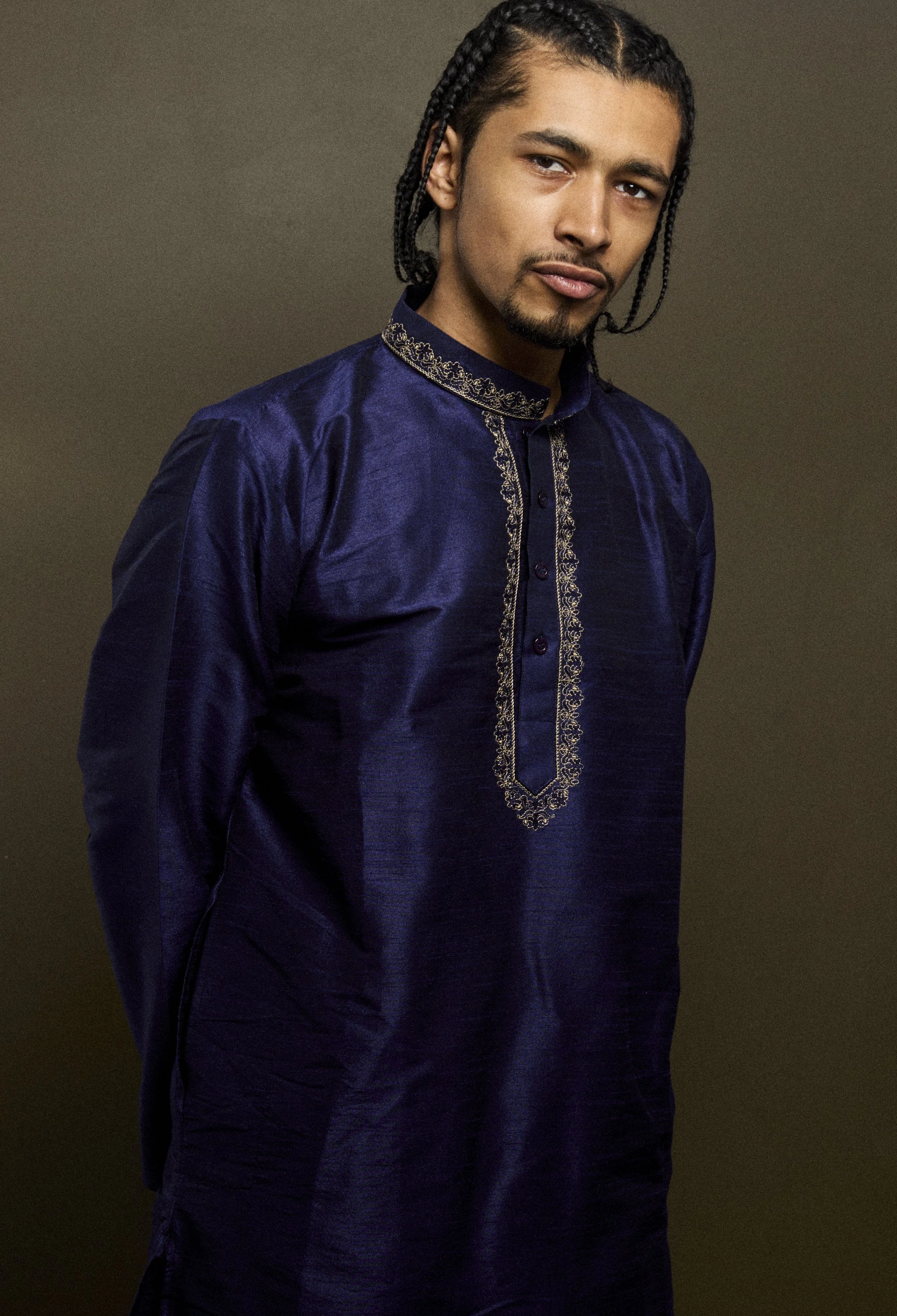 A young man with braids wearing a dark blue traditional African outfit with gold embroidery.