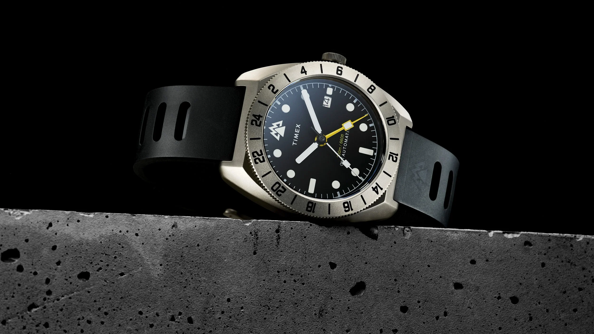 A Timex wristwatch with a black dial, silver bezel, and black rubber strap, resting on a textured concrete surface with a black background.