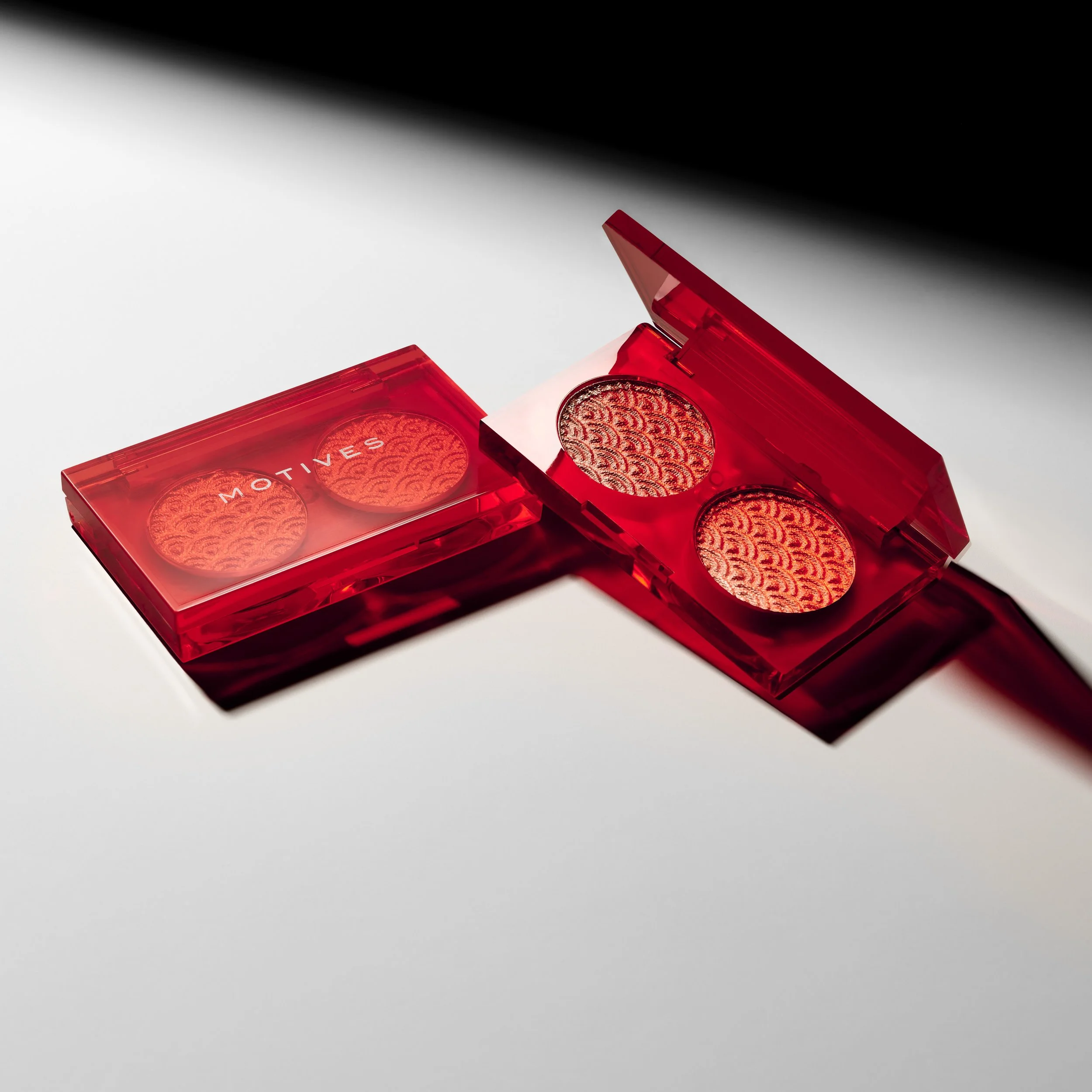 A red compact makeup eyeshadow palette with two circular eyeshadows with a textured pattern inside, one with the word 'motivates' printed on the cover.