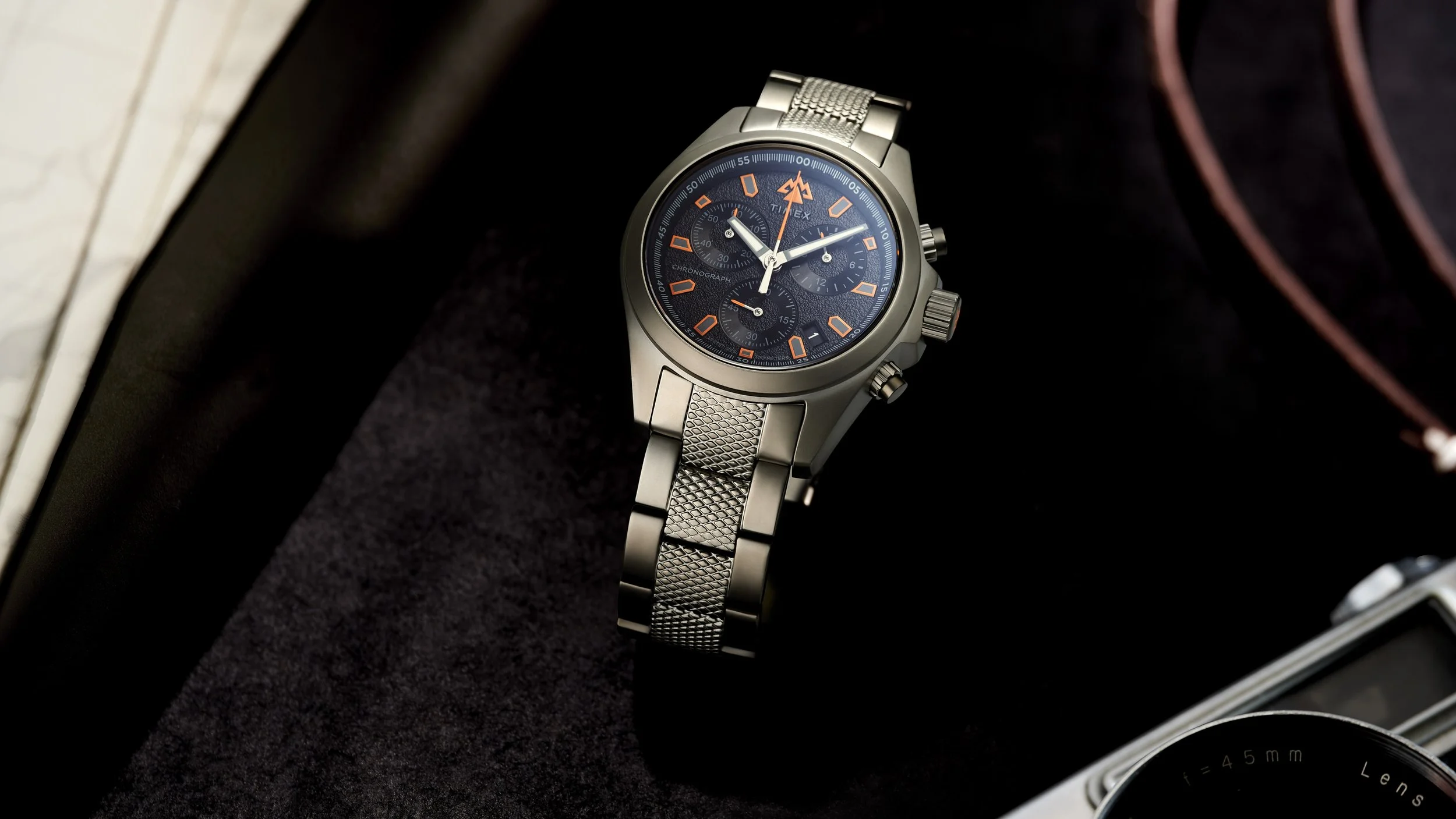 A close-up of a silver men’s chronograph wristwatch with a black dial, orange numerals, and multiple sub-dials, resting on a dark surface with a partial view of a camera and a leather strap to the right.