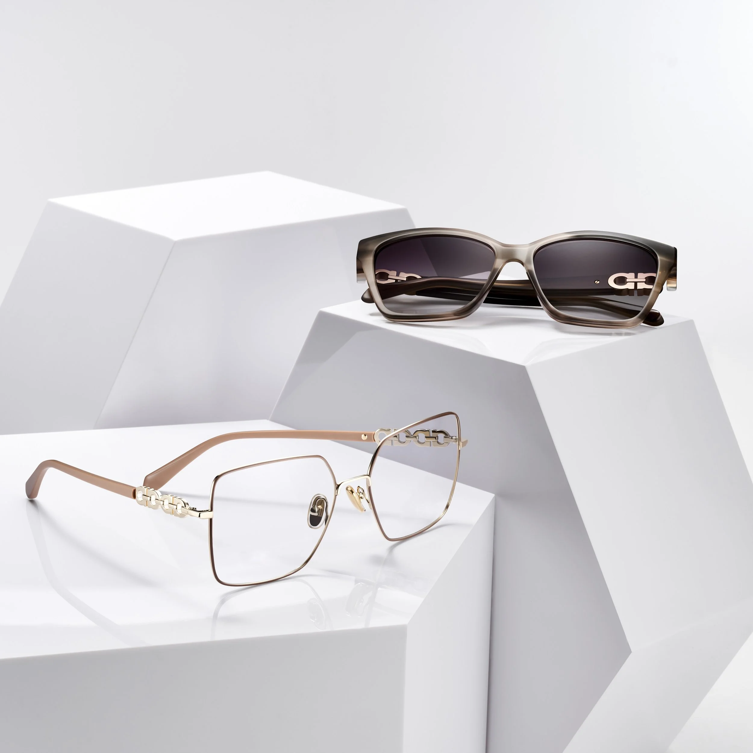 Pair of sunglasses with dark lenses on top of a white geometric display stand, and a pair of gold-rimmed eyeglasses with no lenses on a lower stand.