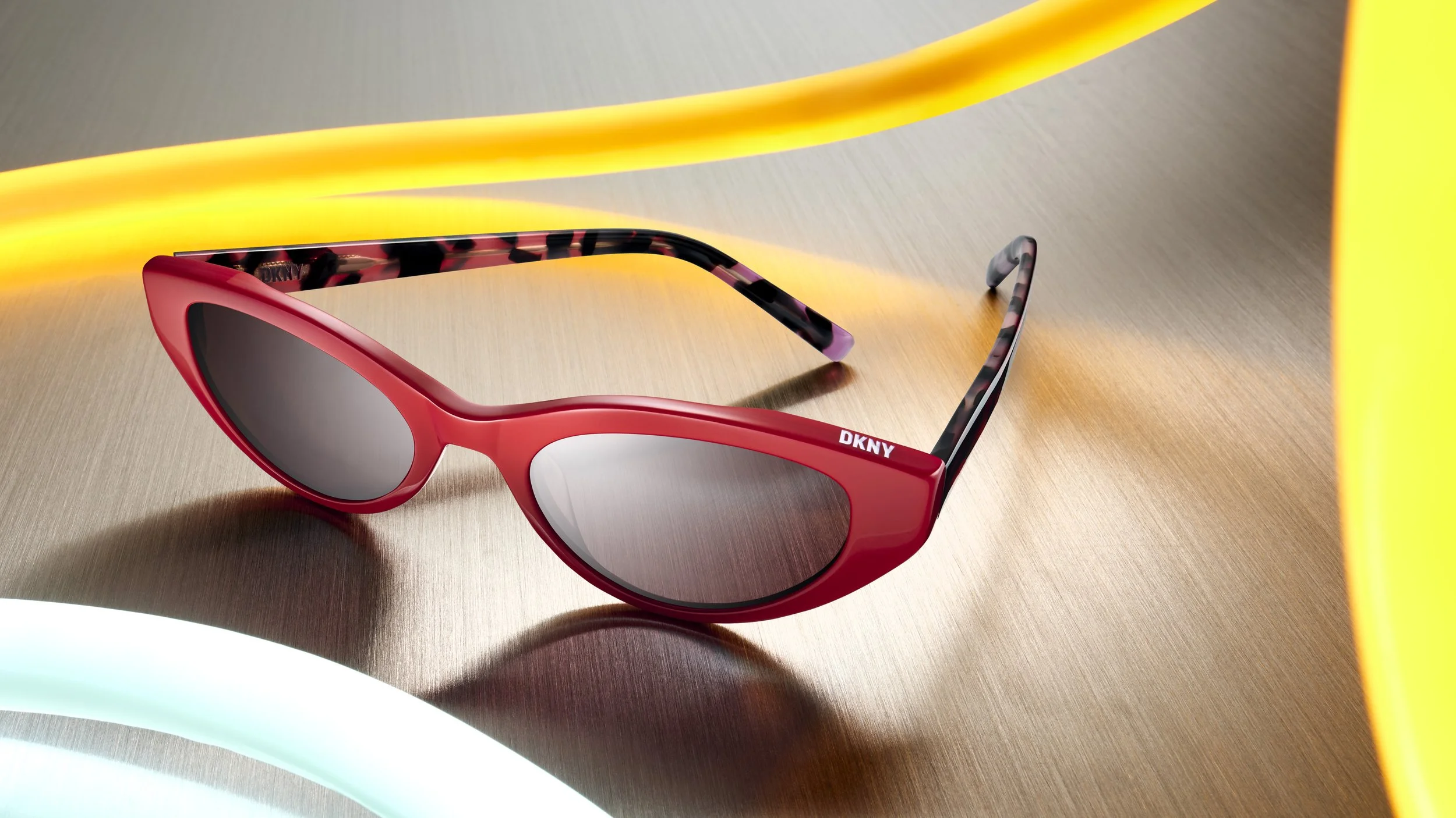 Red DKNY sunglasses with black and pink patterned temples on a wooden surface, with yellow and white neon light accents nearby.