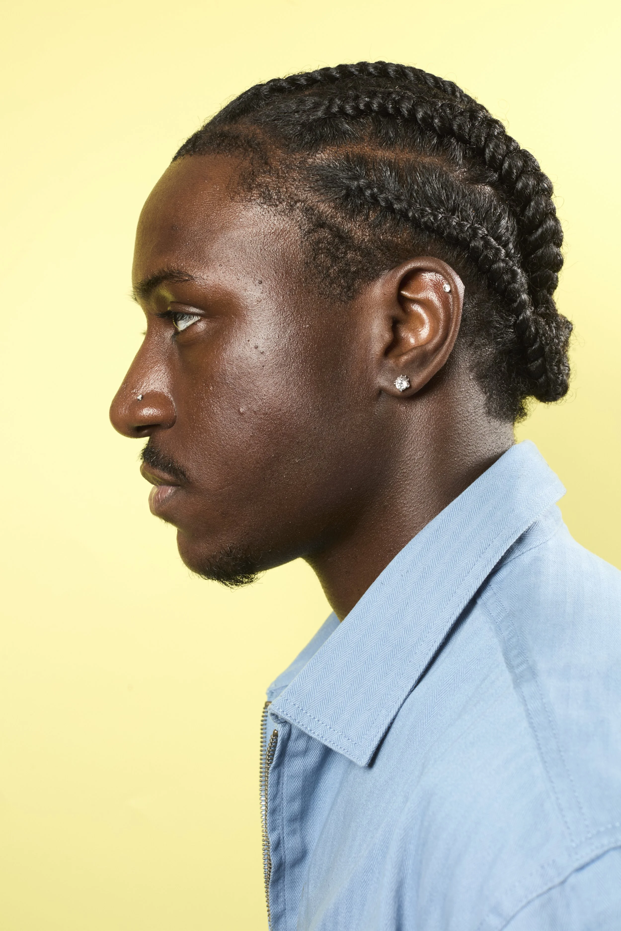 Side profile of a man with dark skin and braided hair, wearing a light blue jacket with a collar, against a yellow background.