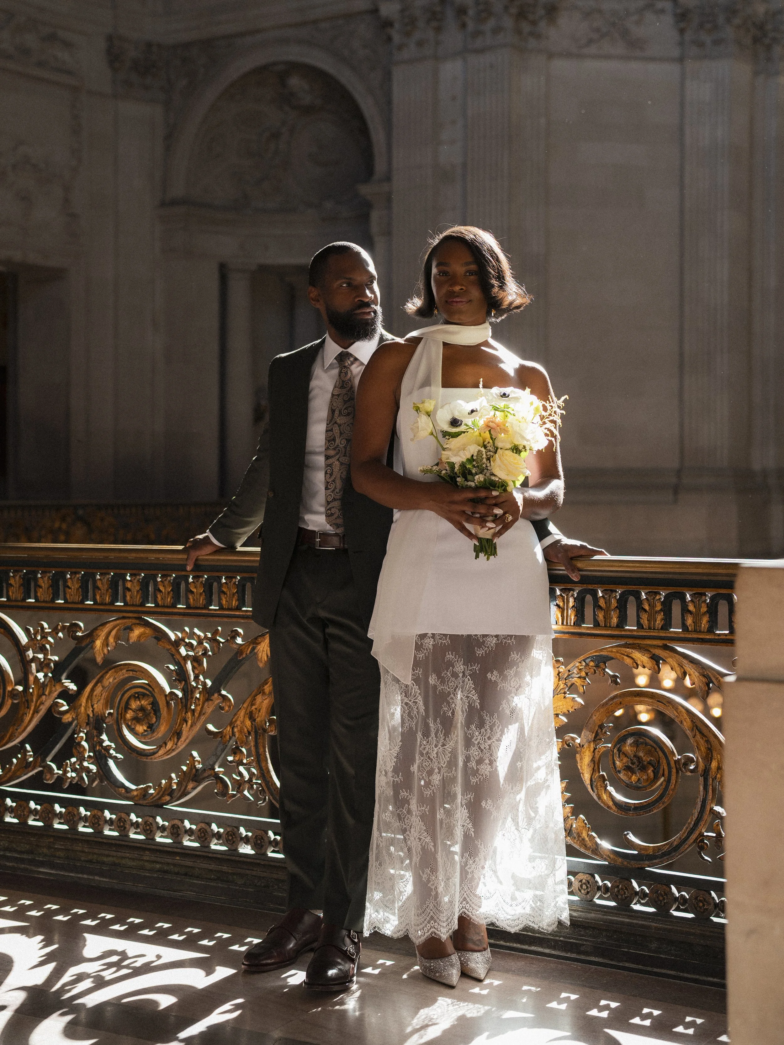 A couple in formal attire standing on a staircase with ornate railing inside a historic building. The woman is holding a bouquet of white and cream flowers, wearing a white lace dress and silver heels. The man is in a dark suit with a patterned tie a