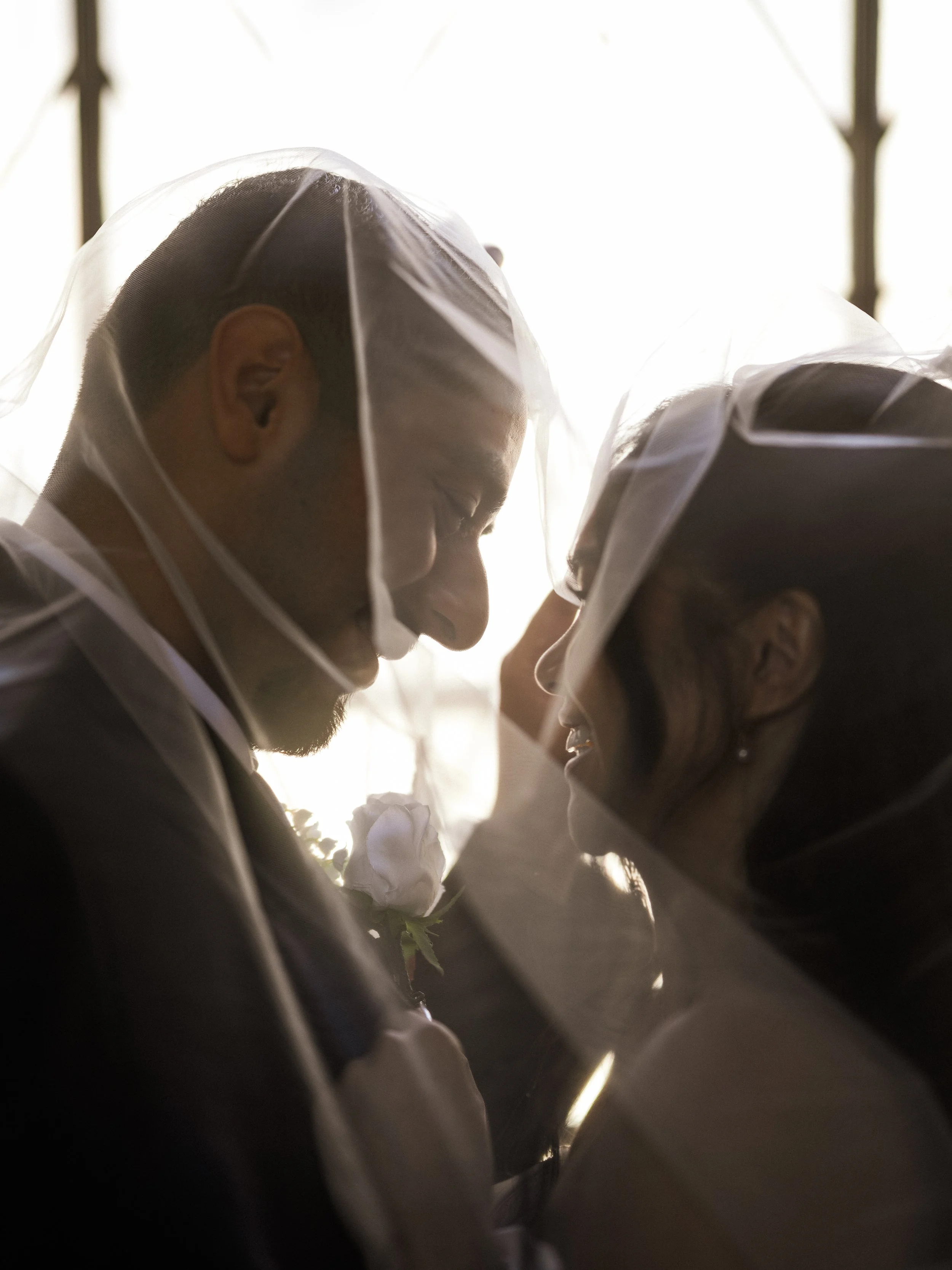 A couple is leaning their foreheads together, smiling, both wearing wedding veils and formal attire, with a white rose boutonniere on the man's suit, during their wedding.