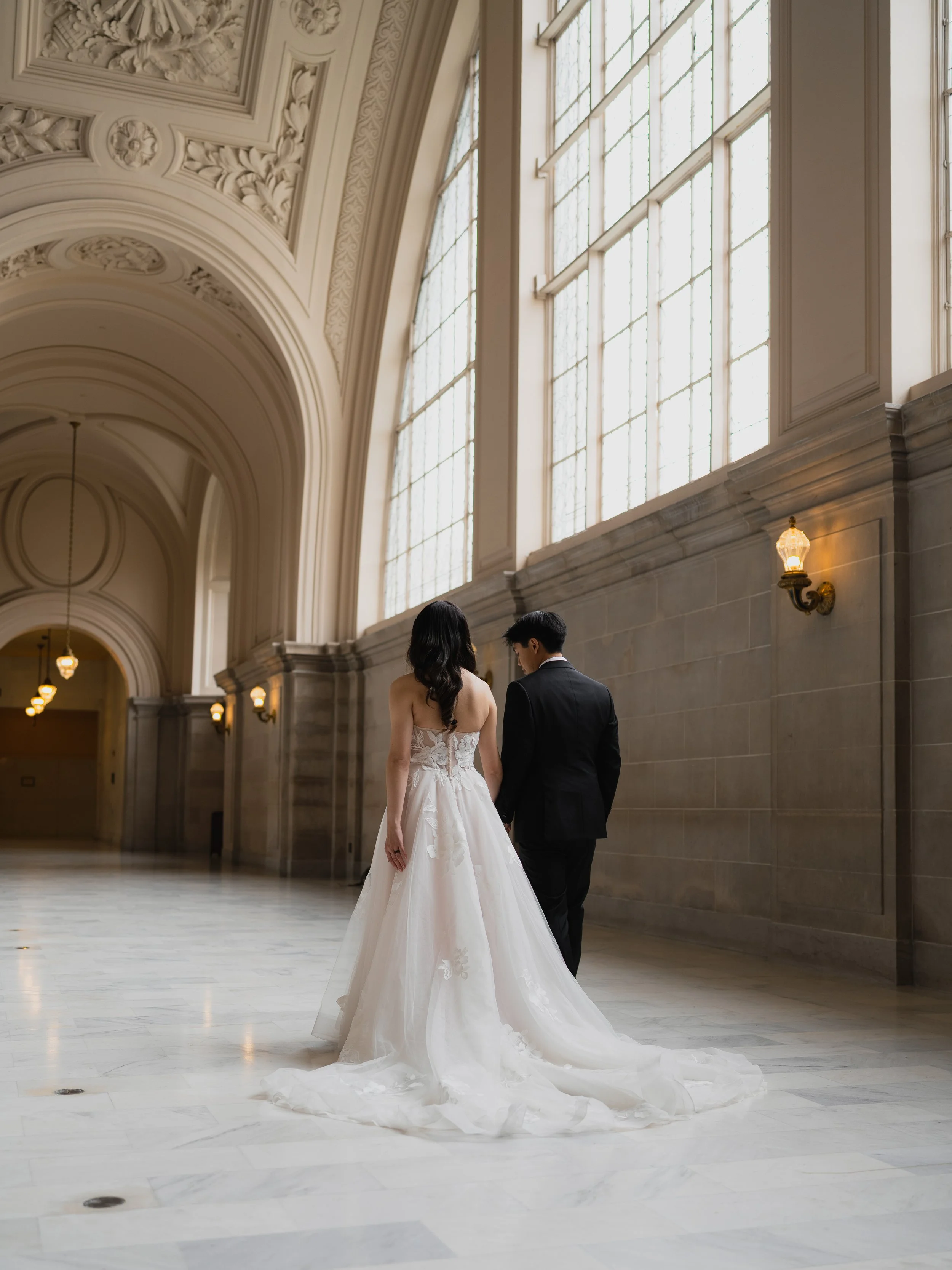 A bride and groom walking hand-in-hand through a grand, historic hall with large windows and ornate ceiling details.