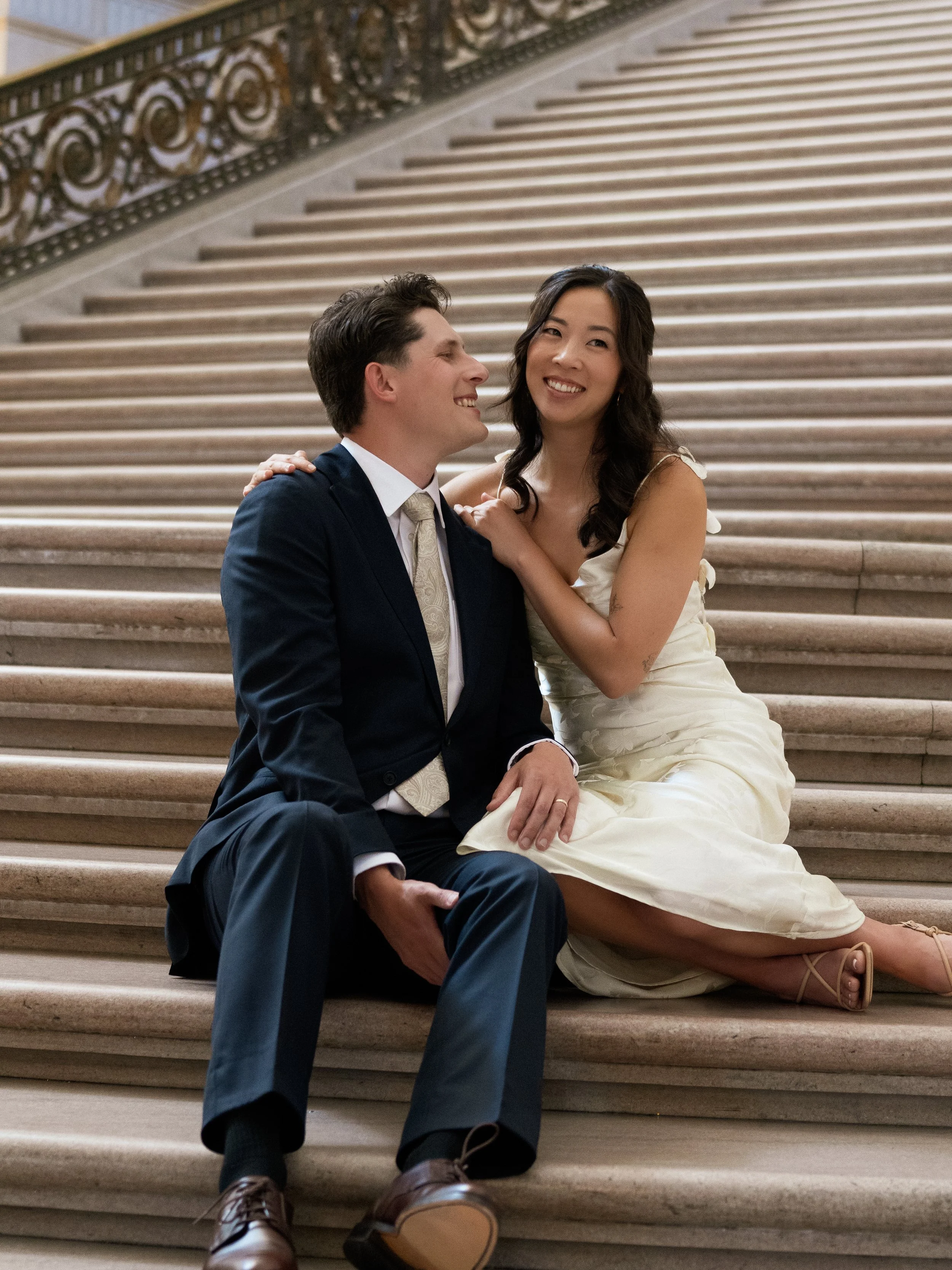 A happy couple sitting together on a staircase, with the man in a dark suit and the woman in a cream-colored dress, smiling at each other.