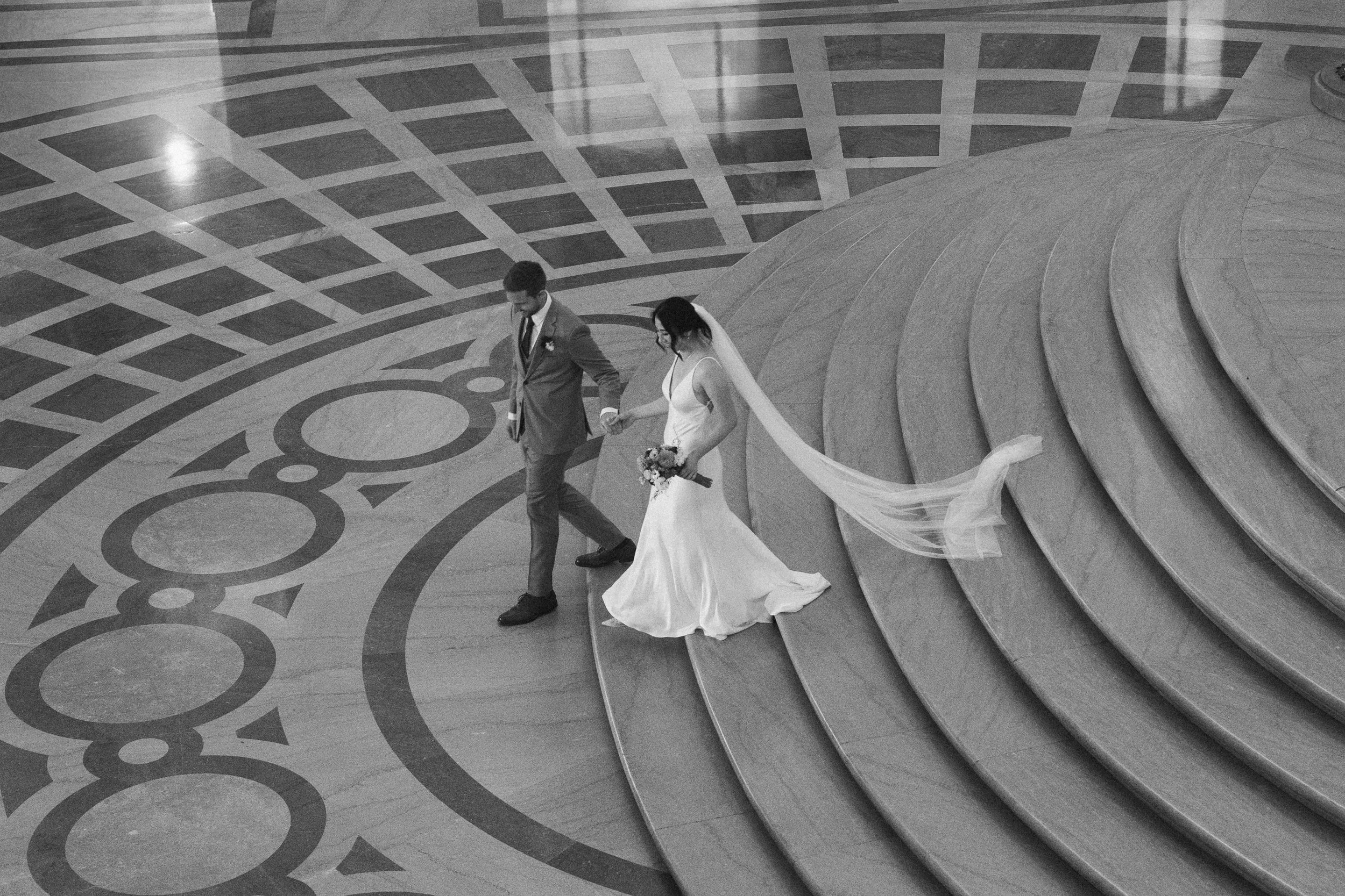 A bride and groom walking down a staircase in an indoor venue with a patterned floor and large open space.