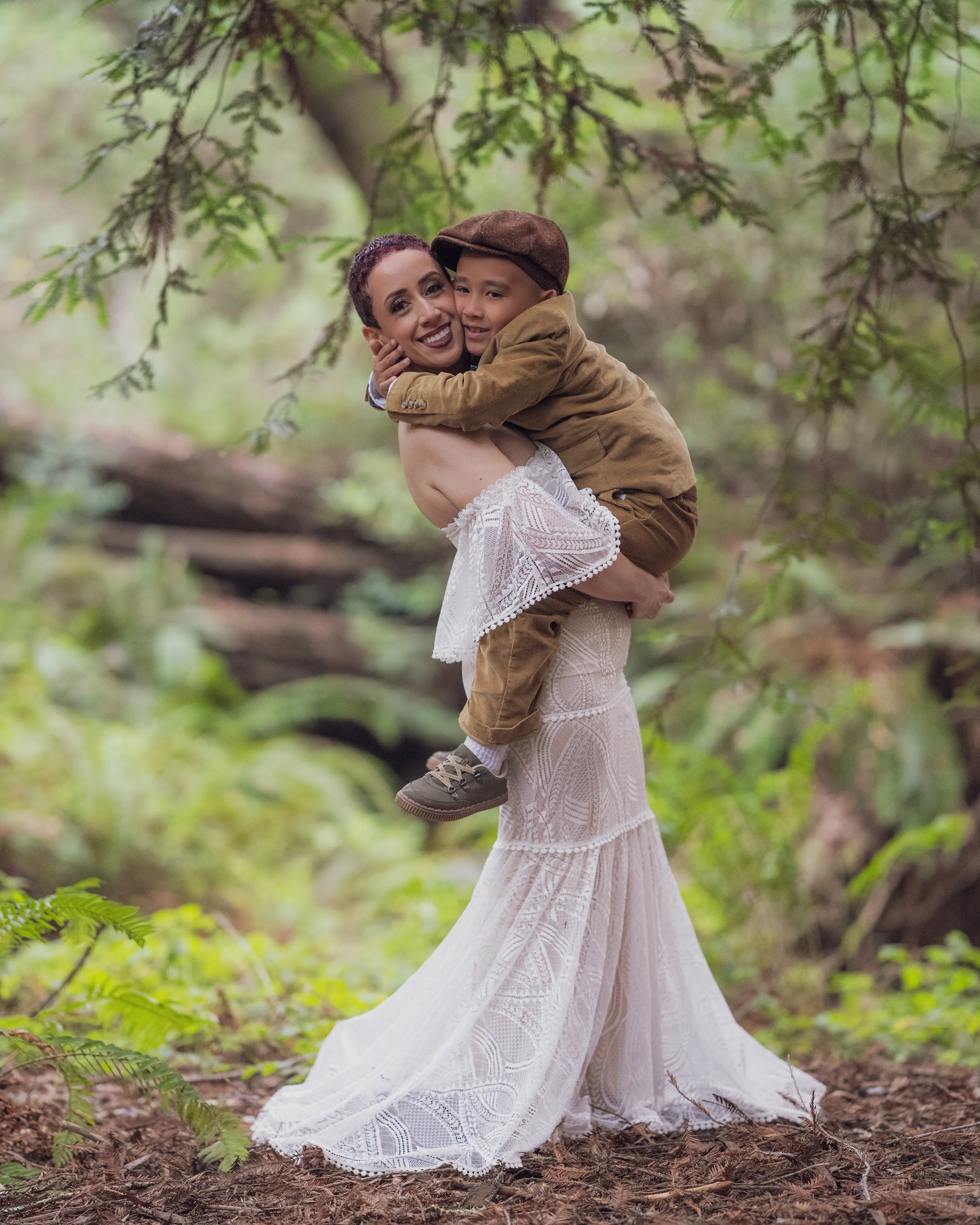 This enchanting photograph captures a heartfelt moment in a lush forest setting. The image showcases a woman in a stunning, intricately designed white lace gown, holding a young child dressed in a charming brown outfit and cap. The natural backdrop o