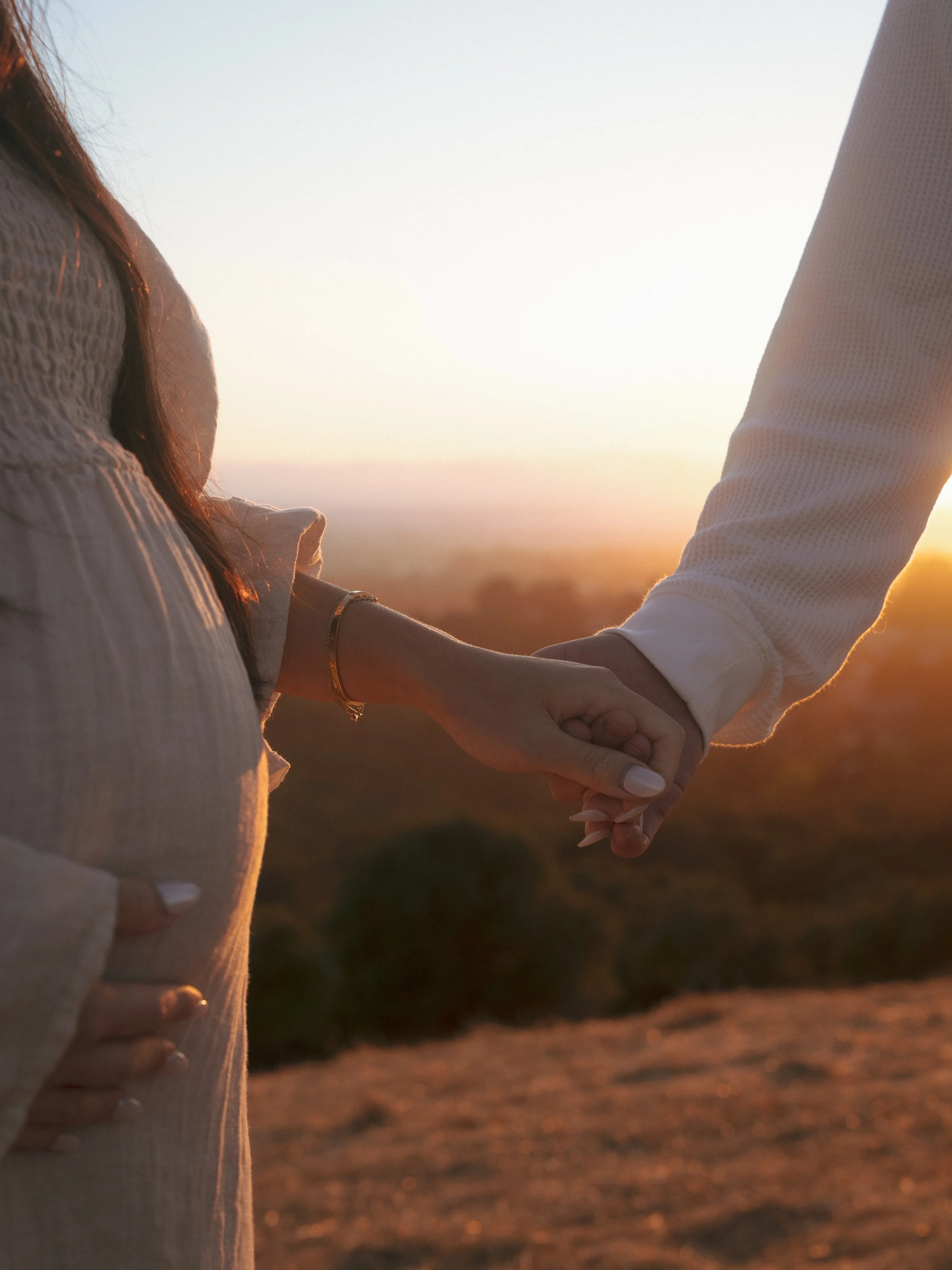 A serene and heartwarming image showcasing a pregnant woman in a flowing, off-the-shoulder maternity dress, capturing the essence of maternity photography and family bonding. The golden sunset backdrop enhances the natural beauty and tranquility of t