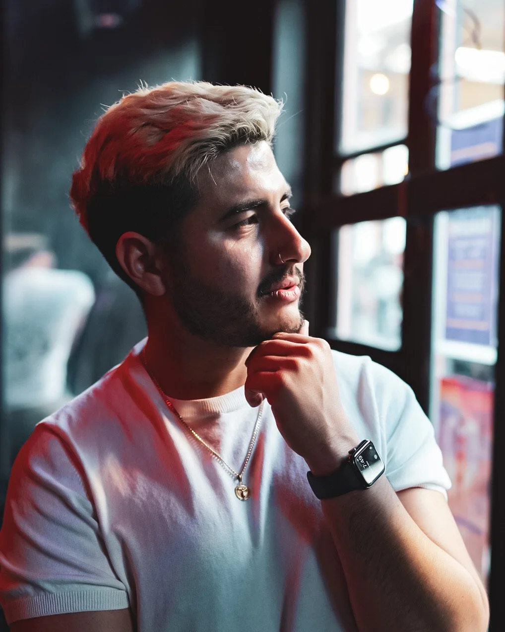 A young man with blond and dark hair, a beard, and a nose ring sitting by a window, looking out pensively. He is wearing a white t-shirt, a necklace, and a smartwatch.