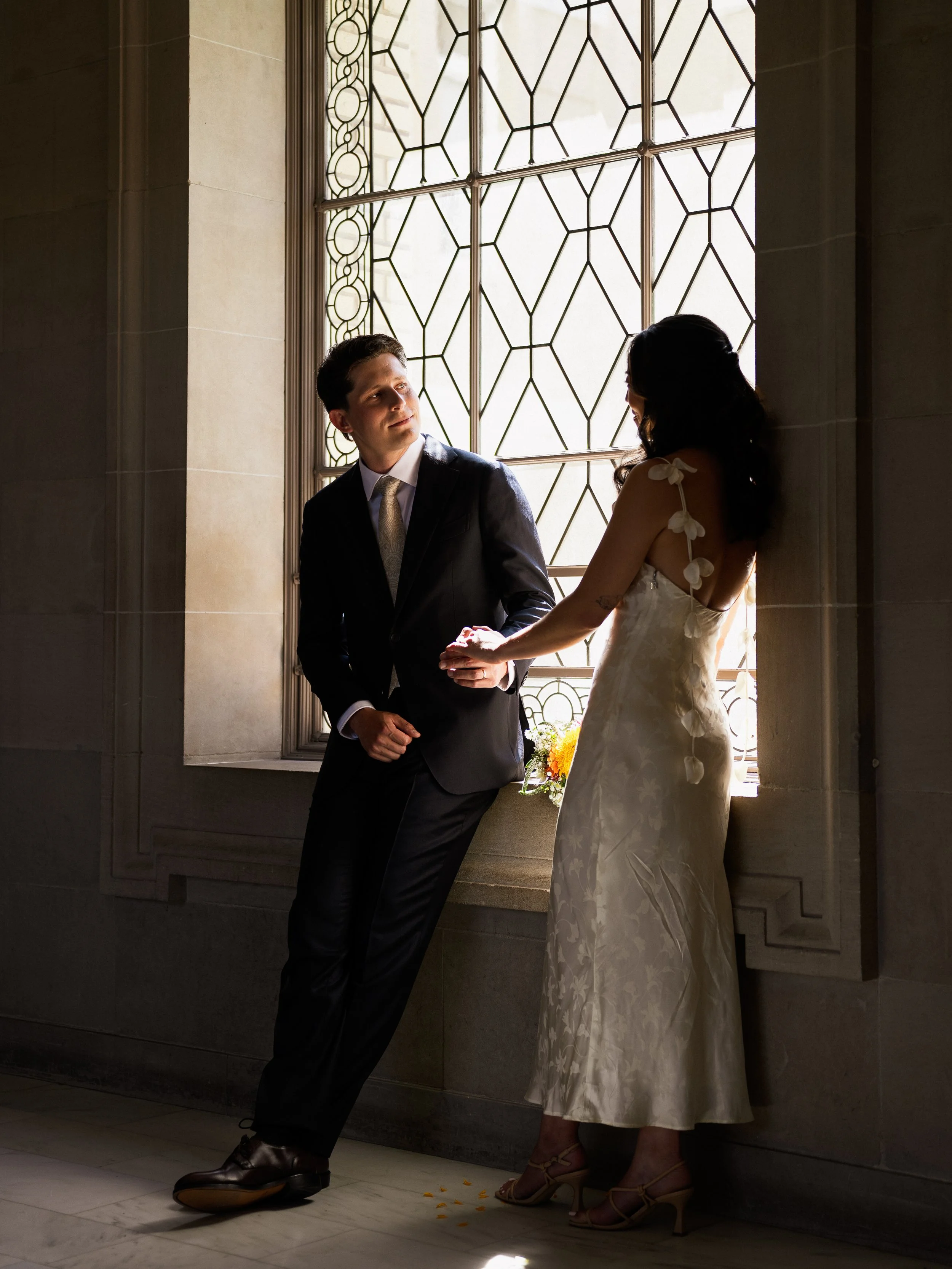 A man in a black suit and woman in a white dress holding hands and standing by a window with intricate metalwork, with sunlight shining through.