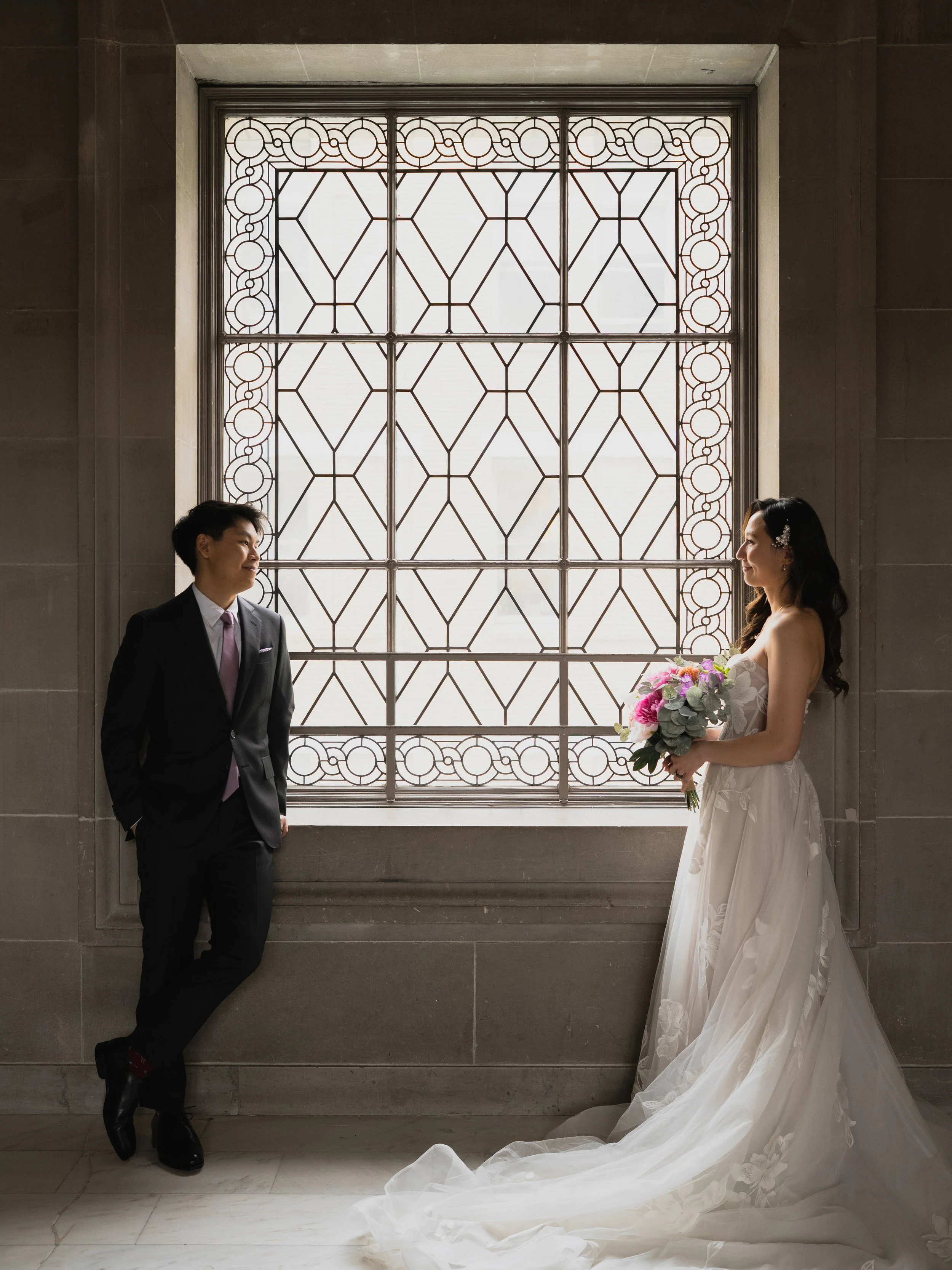 A bride holding a bouquet and a groom in a suit stand facing each other in front of a large window with ornate ironwork in a church or similar venue.
