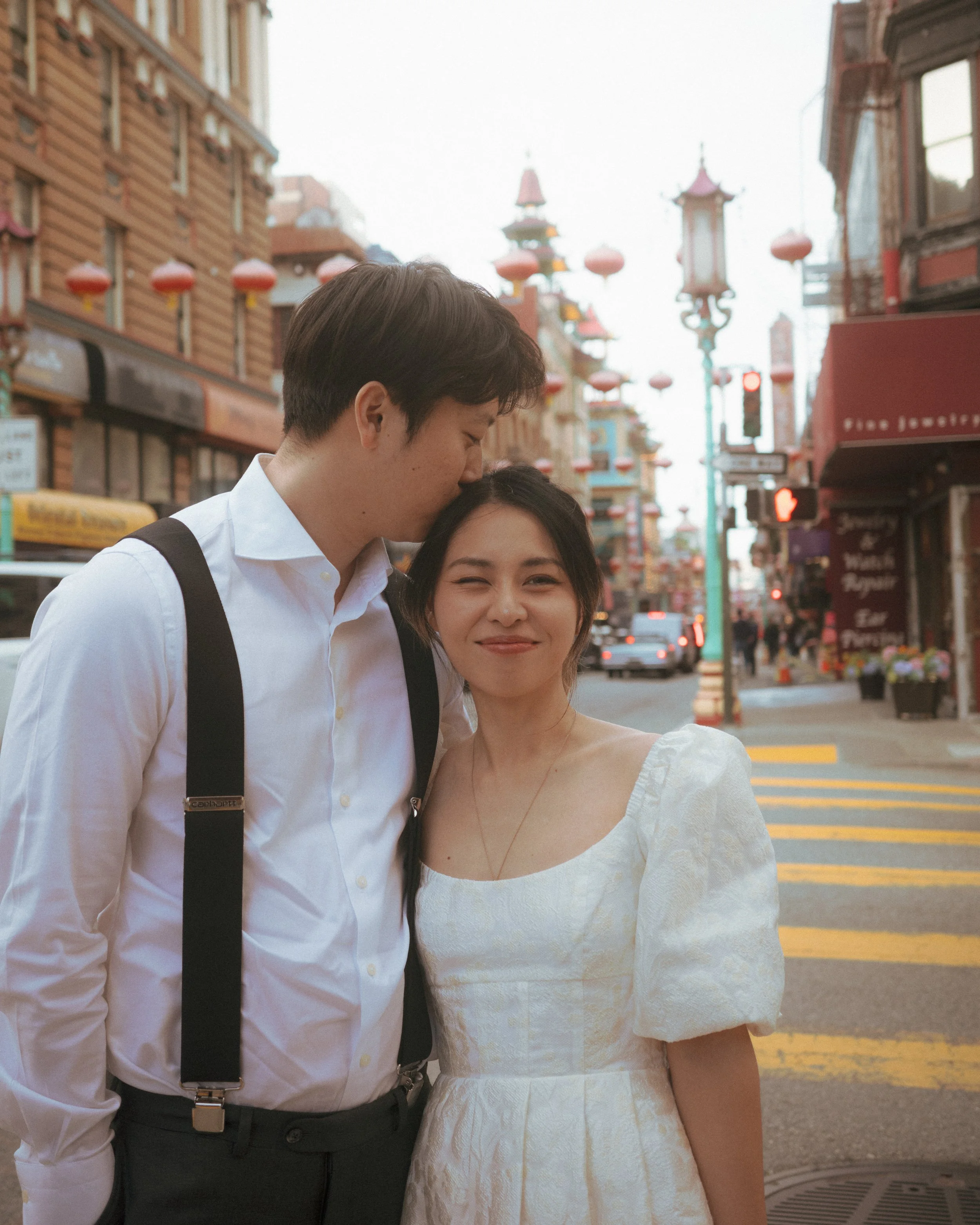 A couple standing close together on a city street, with the man kissing the woman's forehead. The woman is smiling and looking at the camera. City buildings and street lights are visible in the background.