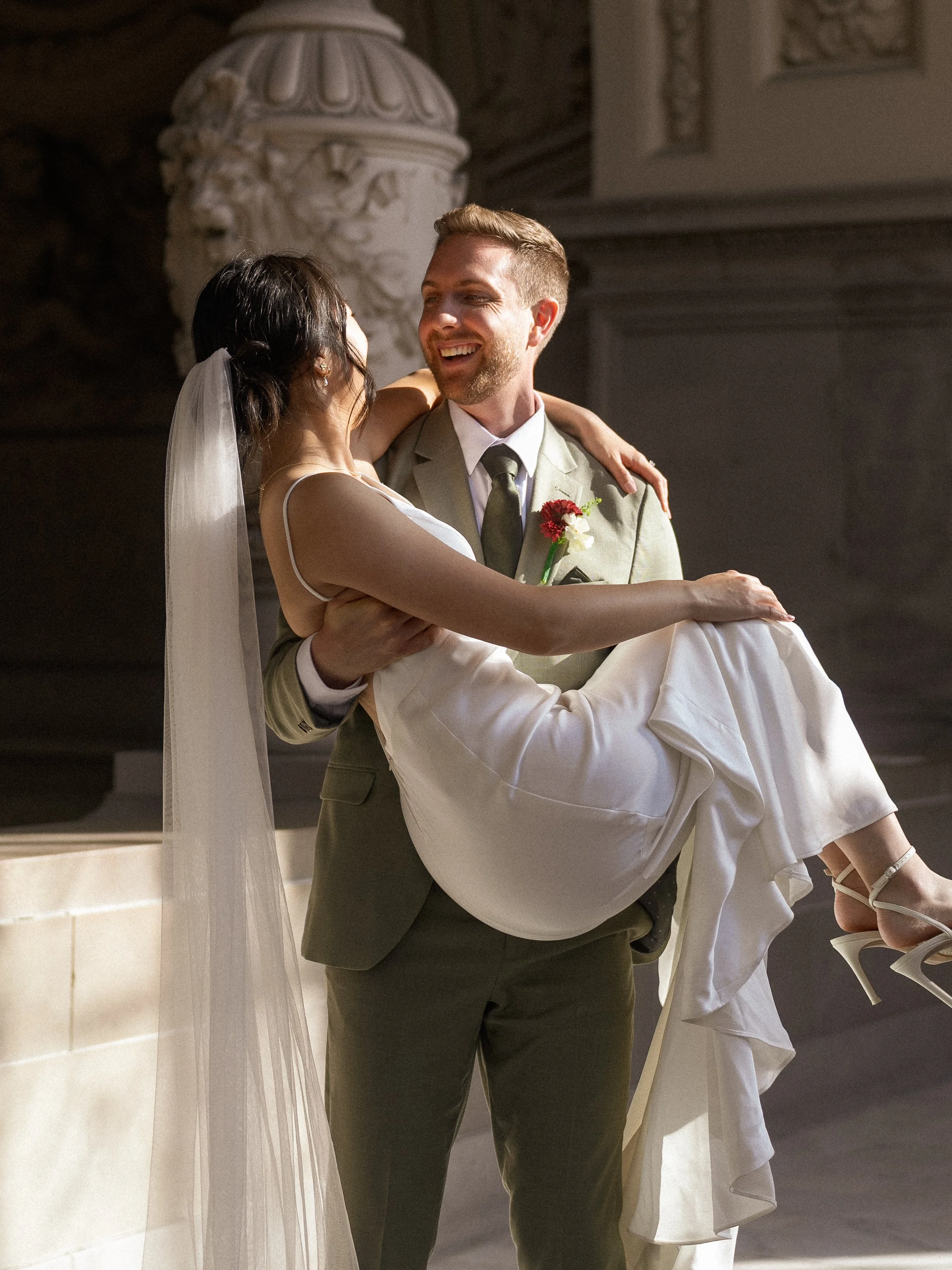 A smiling man in a suit holds a woman in a white dress and veil, lifting her in celebration in an ornate setting.