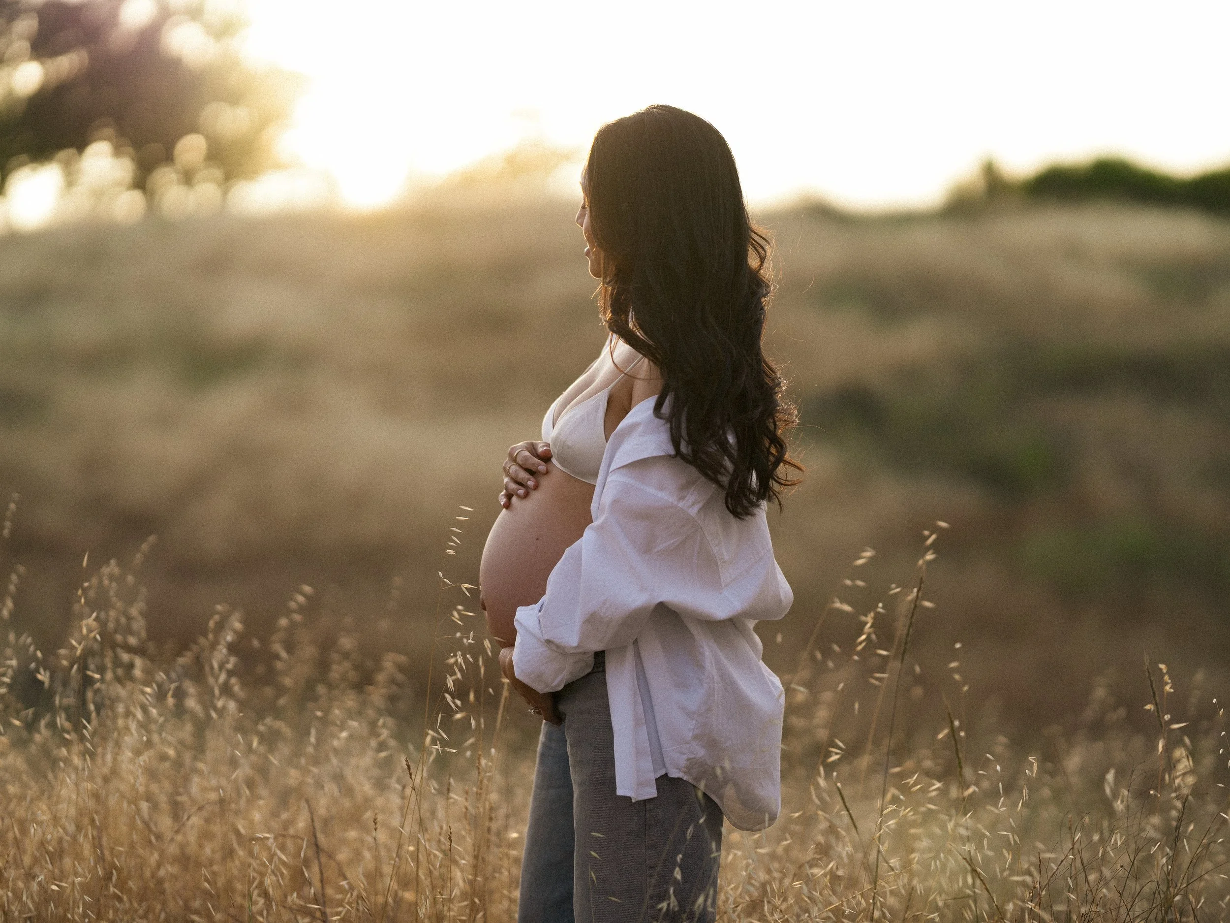 A pregnant woman standing in a field at sunset, with her arms crossed over her belly, wearing a white blouse.