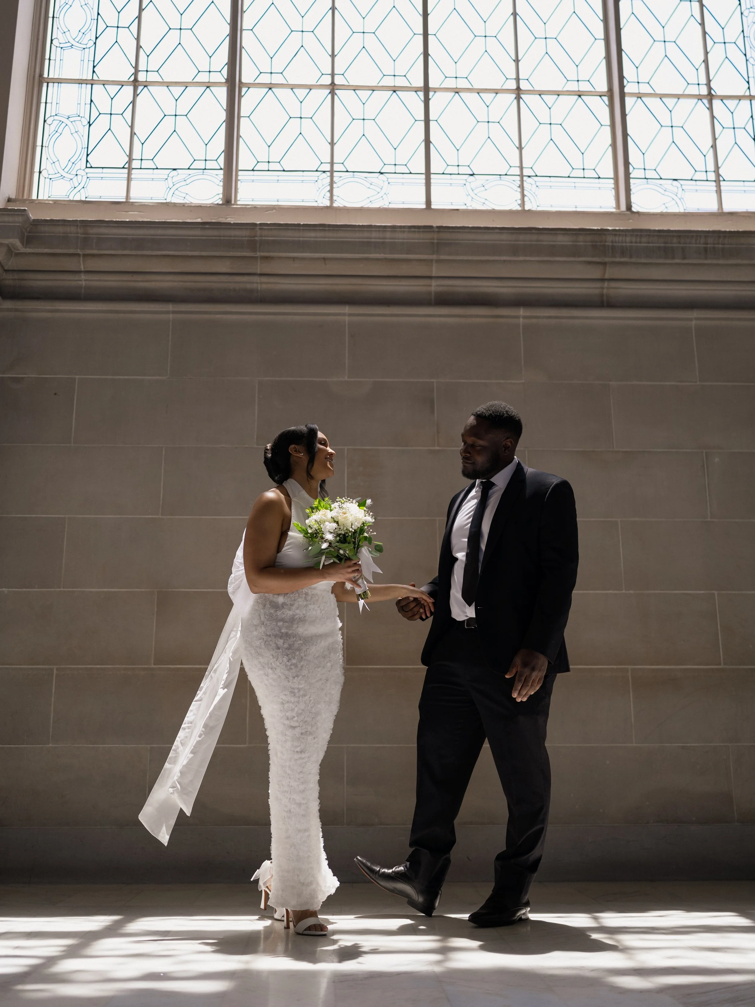 A bride and groom holding hands and smiling at each other inside a building with large window and stone wall.