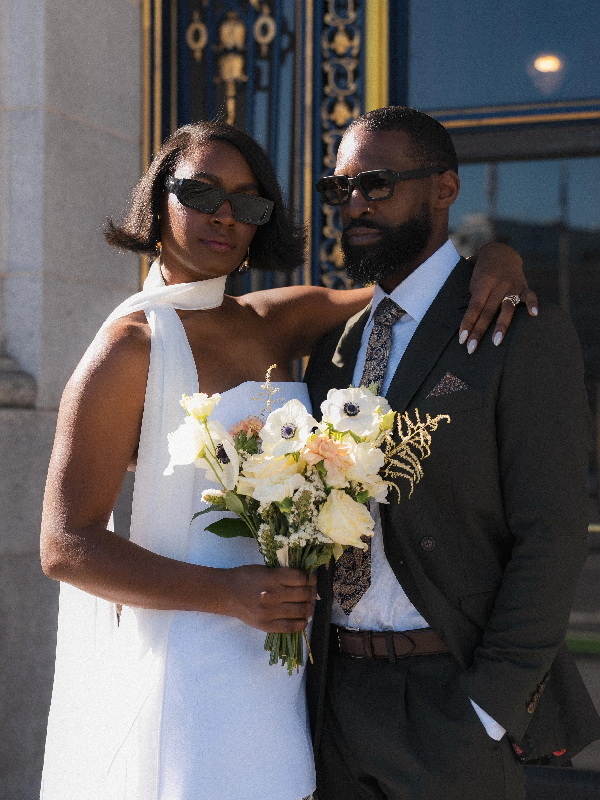 A woman and a man standing close together, both wearing sunglasses, with the woman holding a bouquet of white and pastel flowers. They are dressed elegantly, possibly at a wedding or formal event.