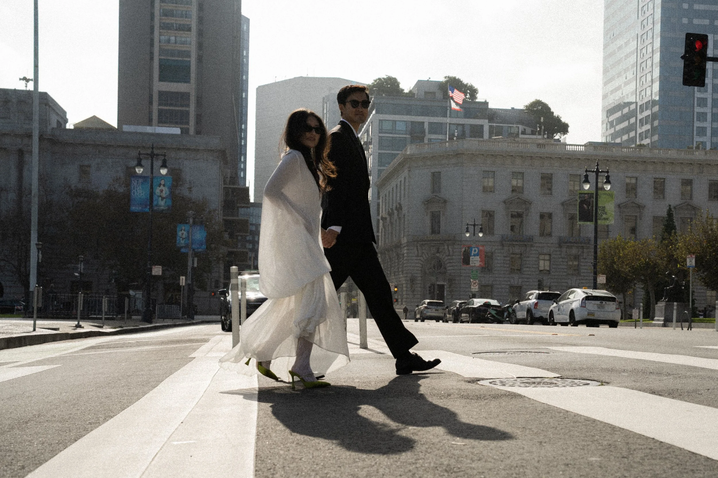 A fashionable woman in a white dress and high heels crossing a city street with a man in a black suit and sunglasses. City buildings and American flags are visible in the background with a traffic light showing red.