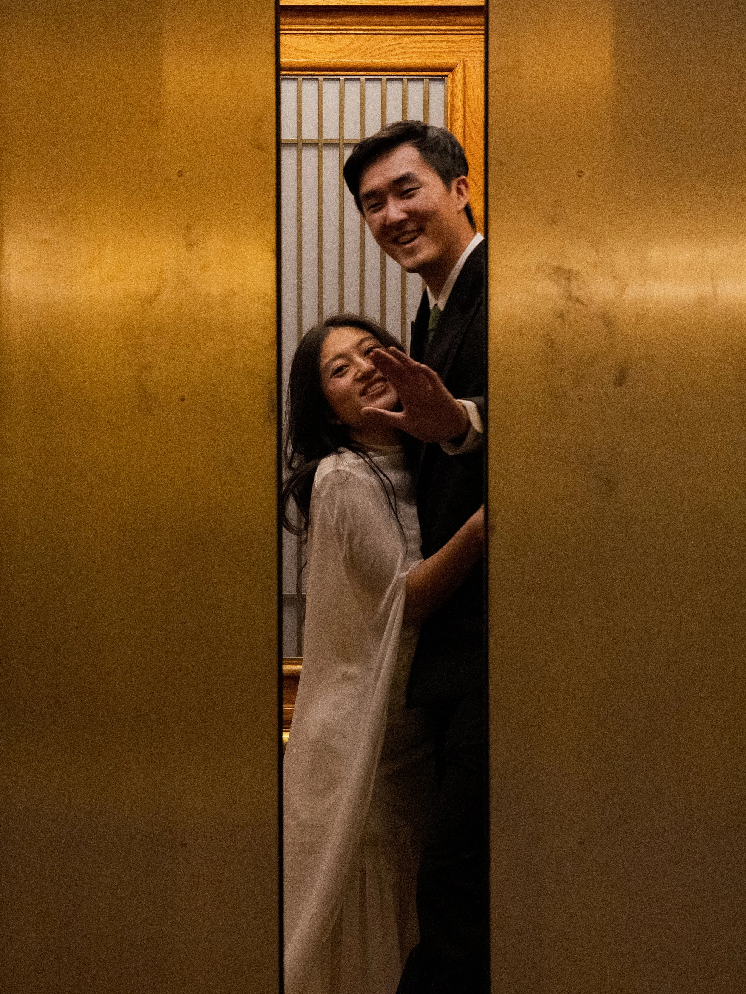 A young man with dark hair and a woman with long, dark hair standing close together in an elevator, smiling and looking at each other, with the man slightly leaning over the woman, who is wearing a white shirt.
