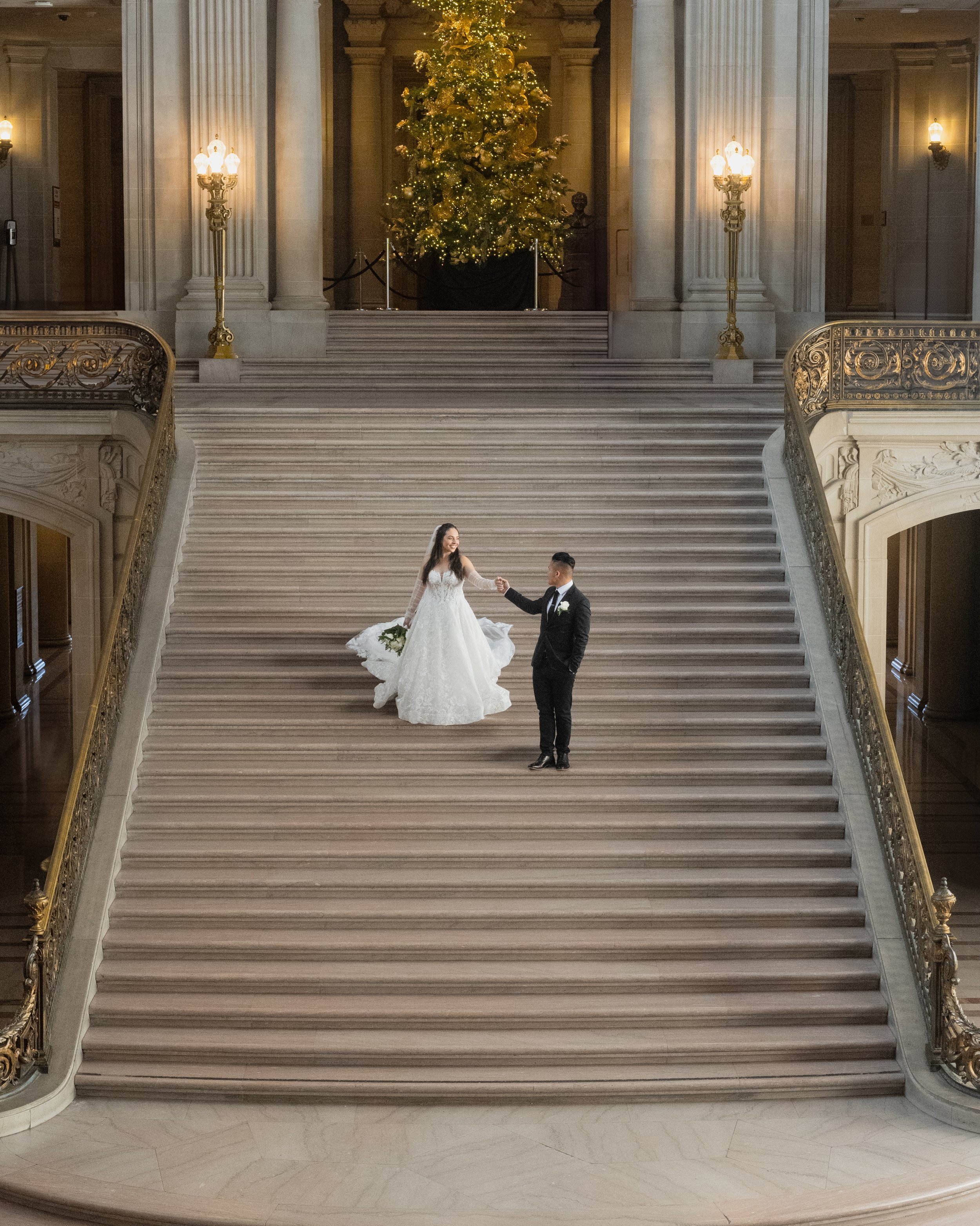 Newlyweds posing on grand staircase with elegant wedding attire, holding hands in front of a beautifully decorated Christmas tree in San Francisco City Hall.