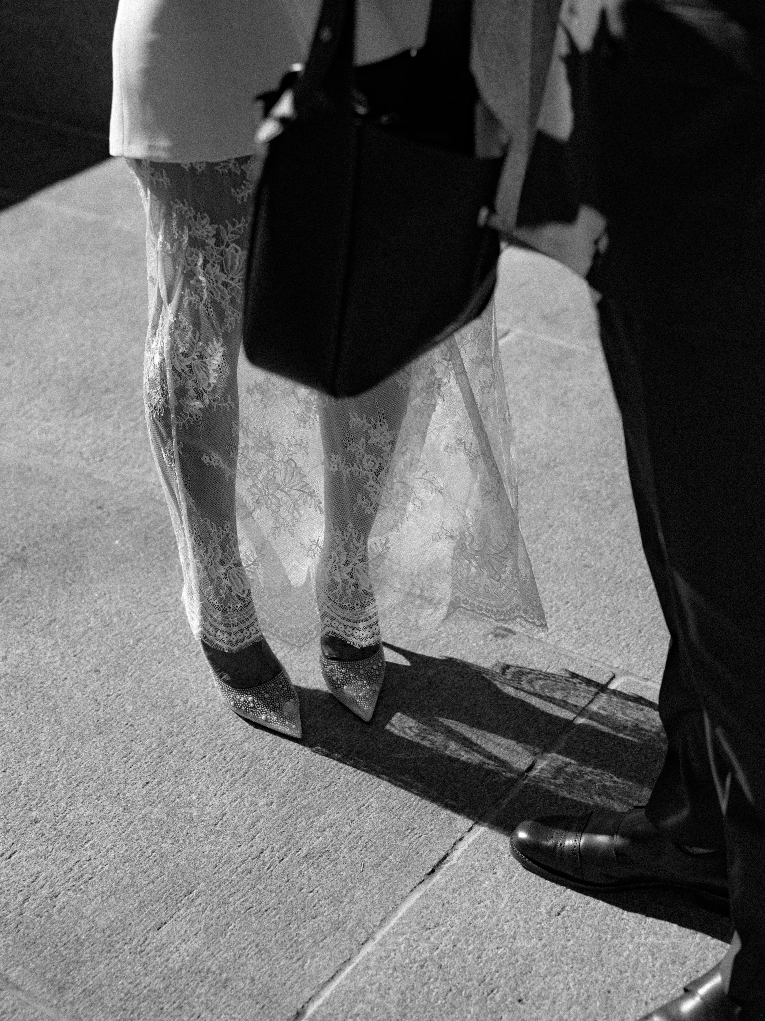 People dressed in formal attire, including a woman in lace dress and a man in a suit, standing on a sidewalk. The focus is on their legs and feet, with one person wearing patterned shoes and the other wearing polished shoes. The photo is in black and