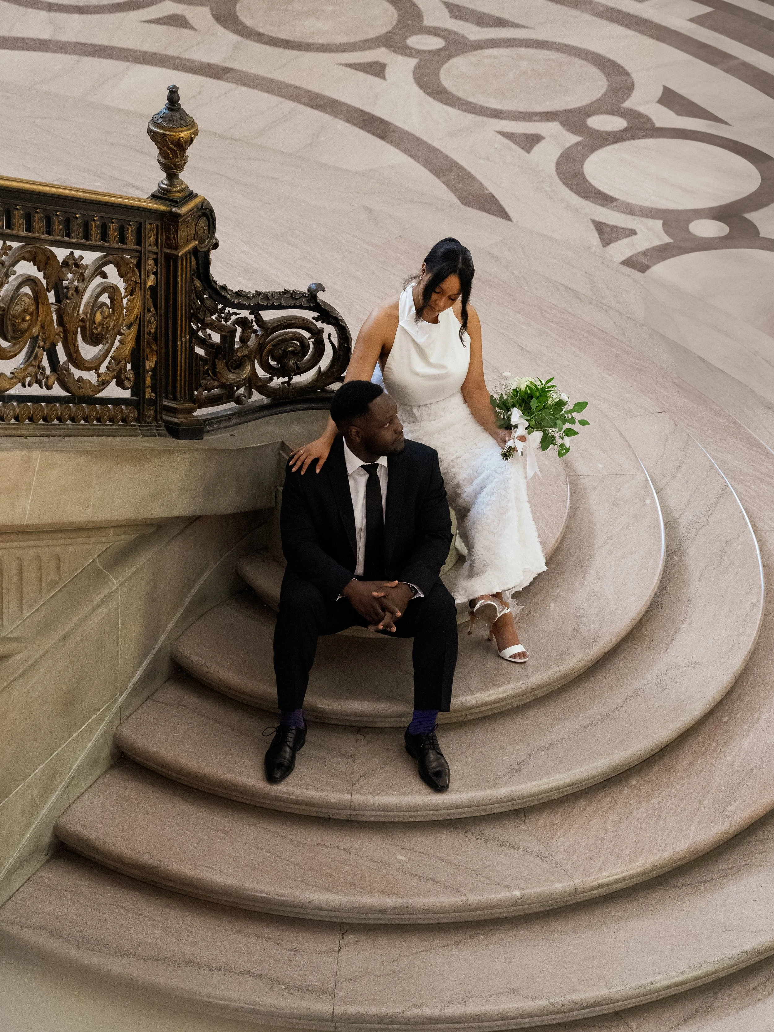 A bride and groom sitting on marble steps inside a grand, ornate building. The bride is holding a bouquet and wearing a white dress, while the groom is dressed in a black suit with a tie.