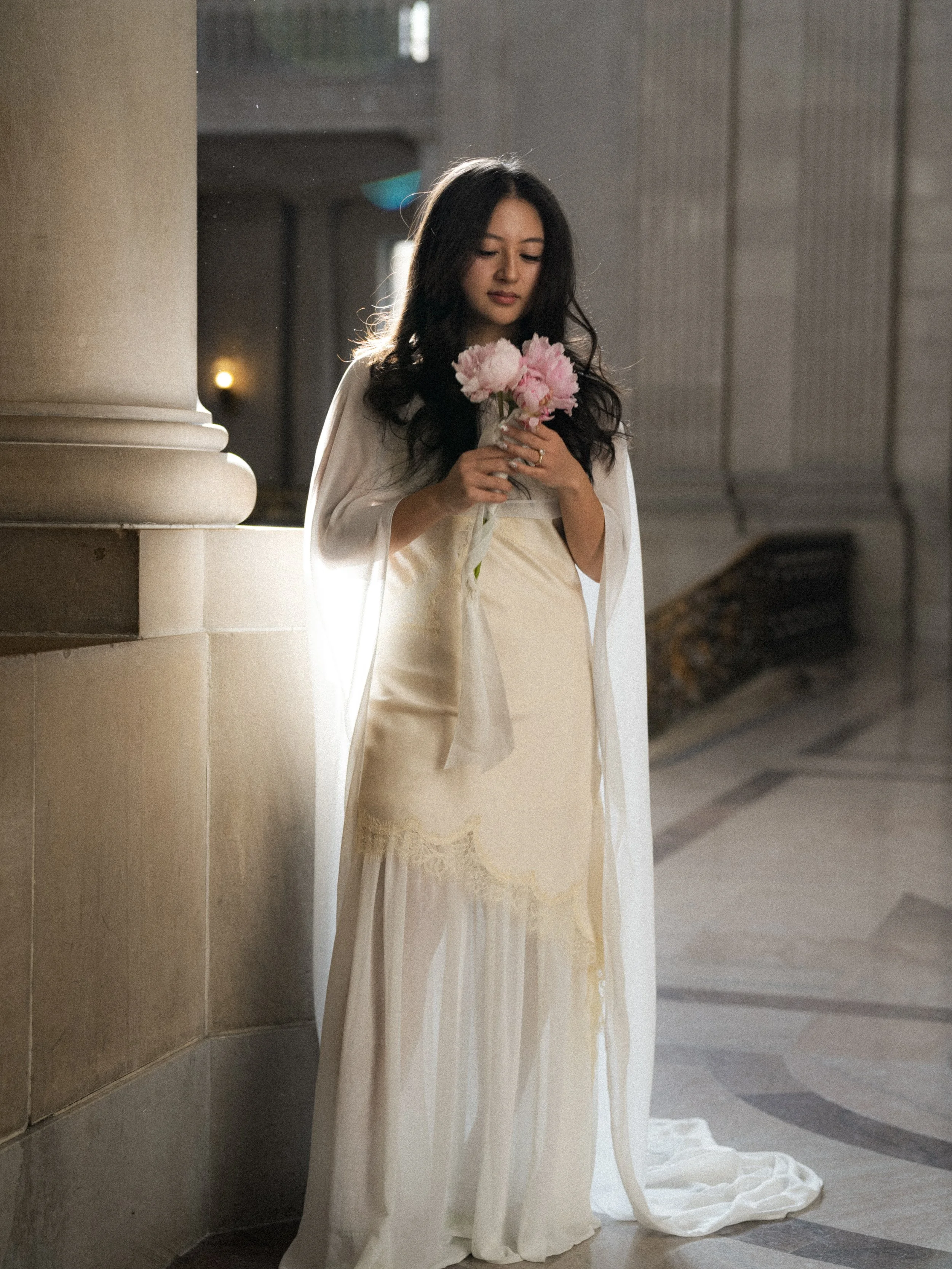 A woman with long dark hair in a white gown holding a bouquet of pink flowers, standing in a building with marble flooring and large columns.