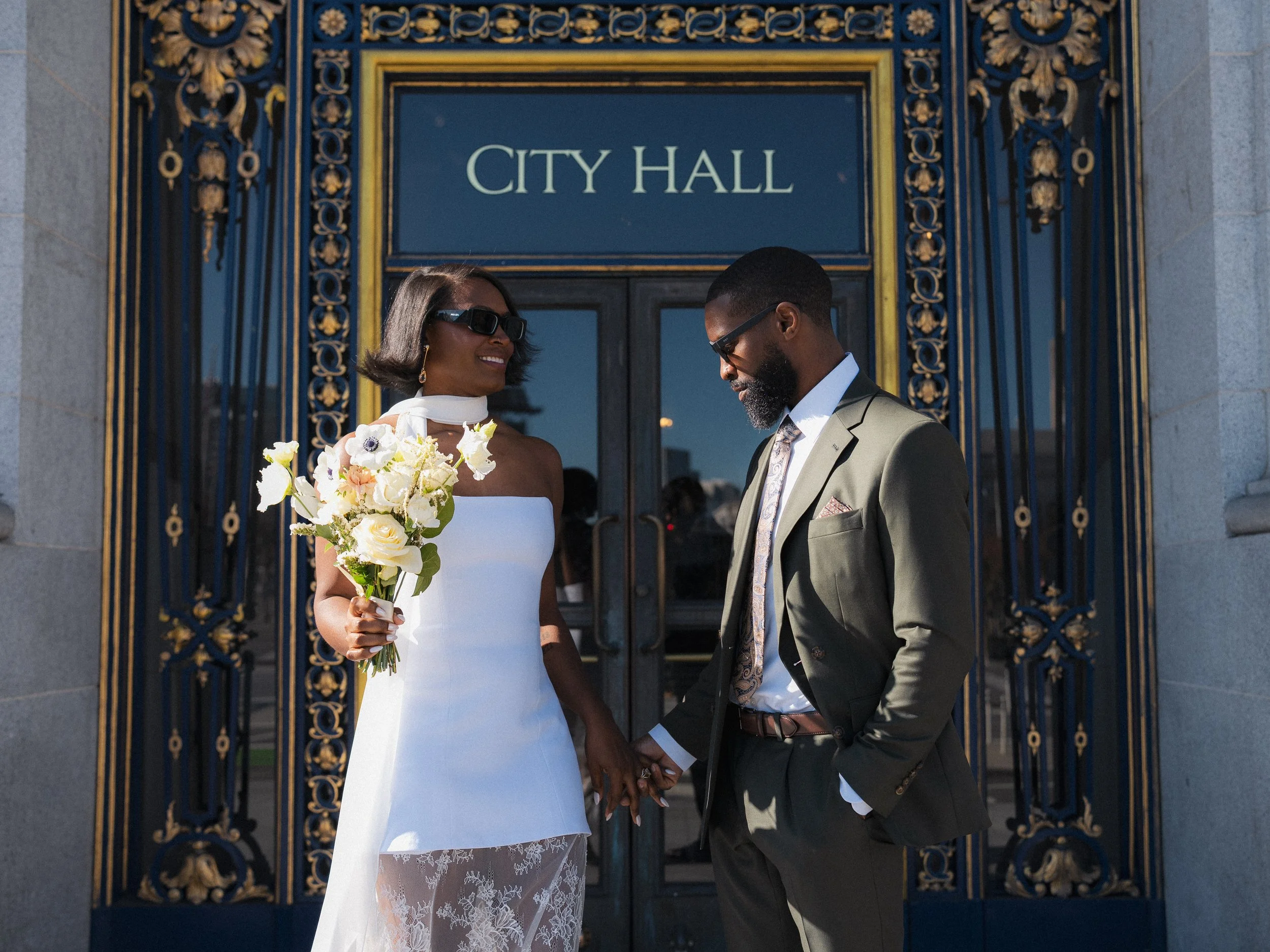 A bride and groom holding hands outside a city hall, smiling at each other. The bride is wearing a white dress with lace detail and holding a bouquet of white and yellow flowers, while the groom is dressed in a suit with a patterned tie. The city hal