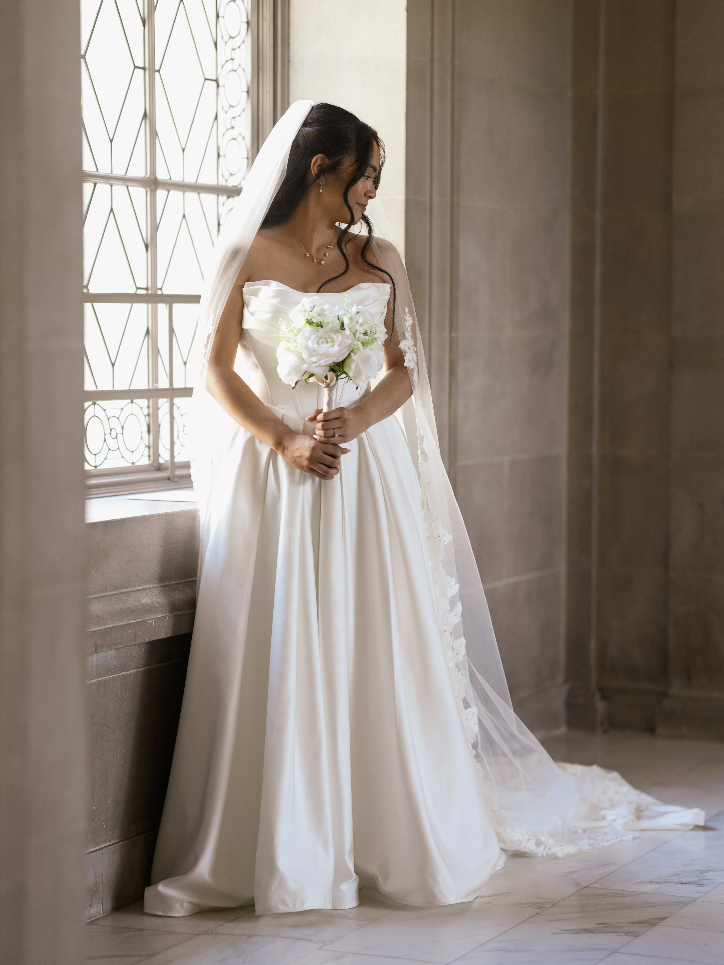 A bride in a white strapless wedding gown holding a bouquet of white flowers, standing indoors near a large window with decorative ironwork, with her head slightly bowed and a gentle smile.