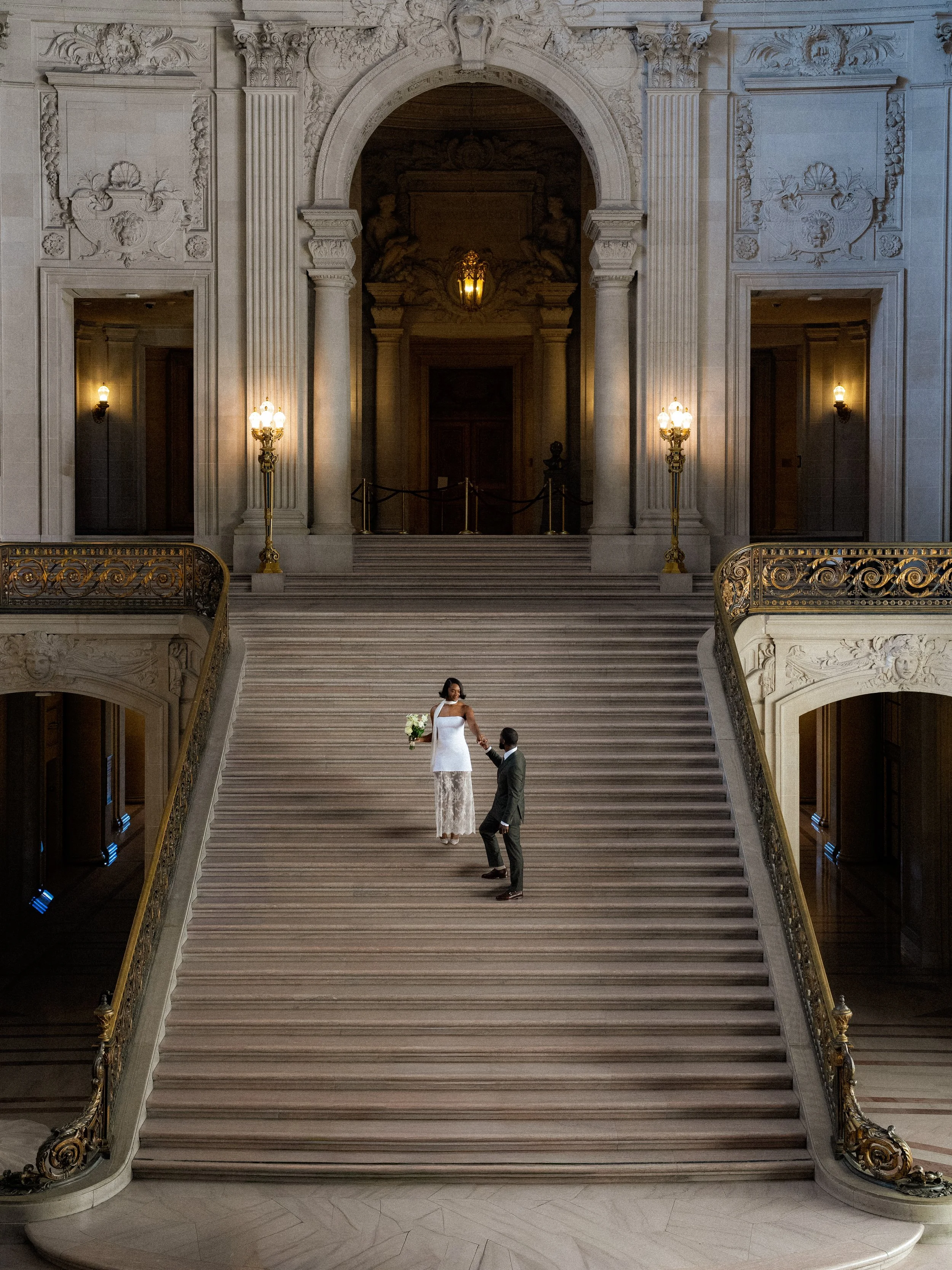 A bride in a white dress holding a bouquet and a groom in a suit standing on a grand marble staircase inside an elegant building with ornate architectural details and warm lighting fixtures.