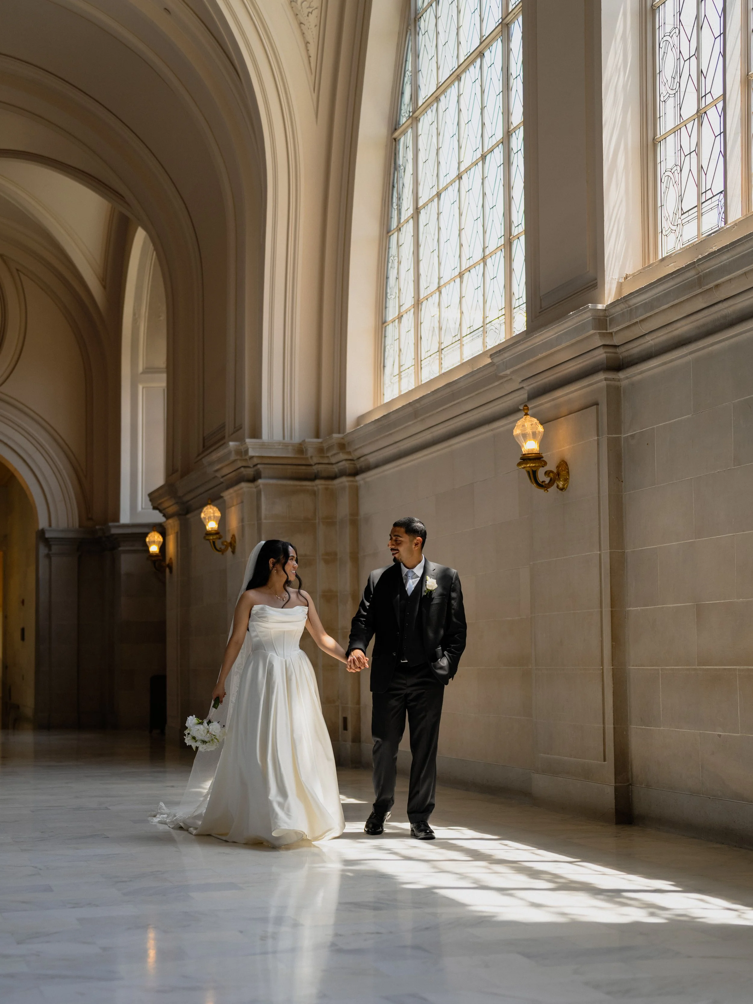 A bride and groom walking hand in hand inside a grand, well-lit hall with large stained glass windows, marble floors, and ornate wall sconces.