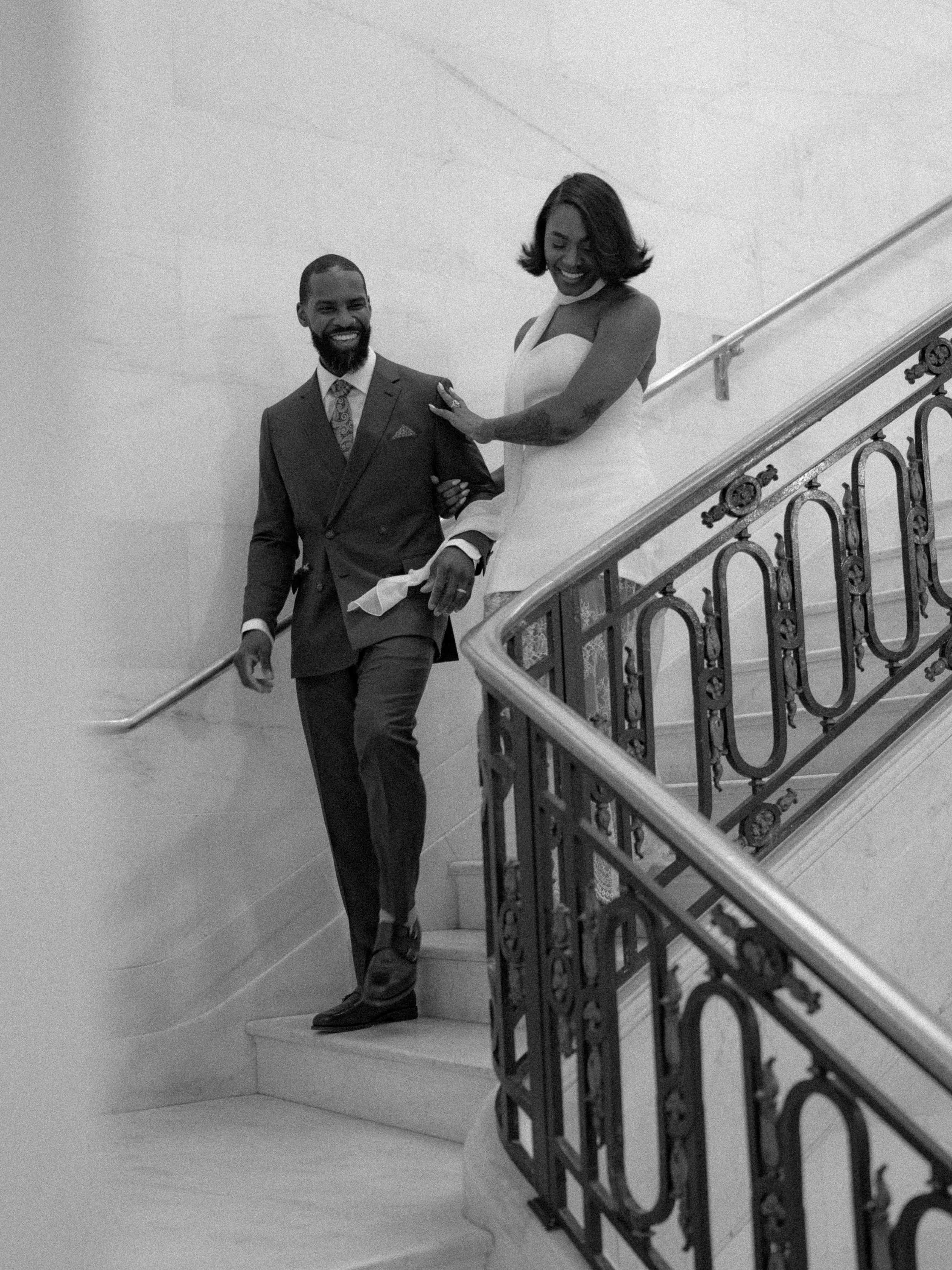 A black-and-white photo of a couple standing on a staircase inside a building. The man is dressed in a suit and tie, smiling and holding onto the woman's hand. The woman is wearing a white dress, also smiling, with her hand on the man's shoulder. The