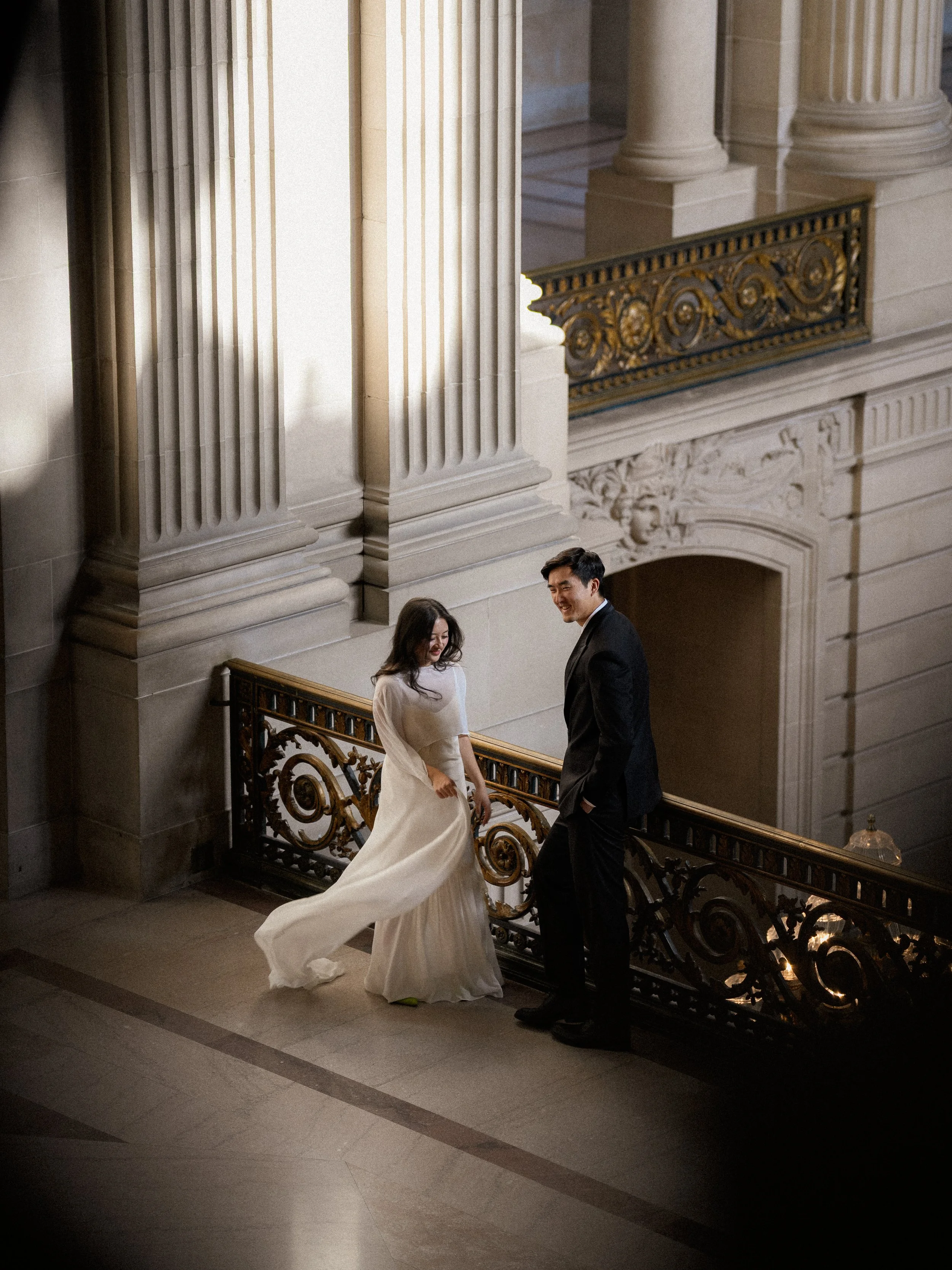 A bride and groom standing on a staircase in a grand building, smiling at each other.