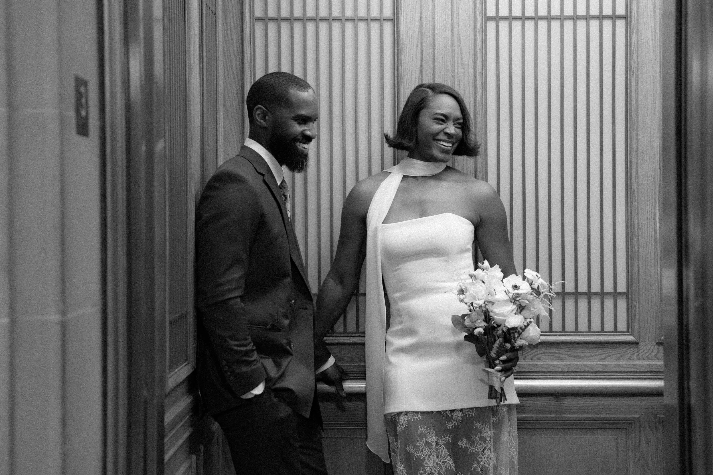 A black and white photo of a couple in an elevator, with the man in a suit on the left and the woman in a dress holding a bouquet of flowers on the right, both smiling.