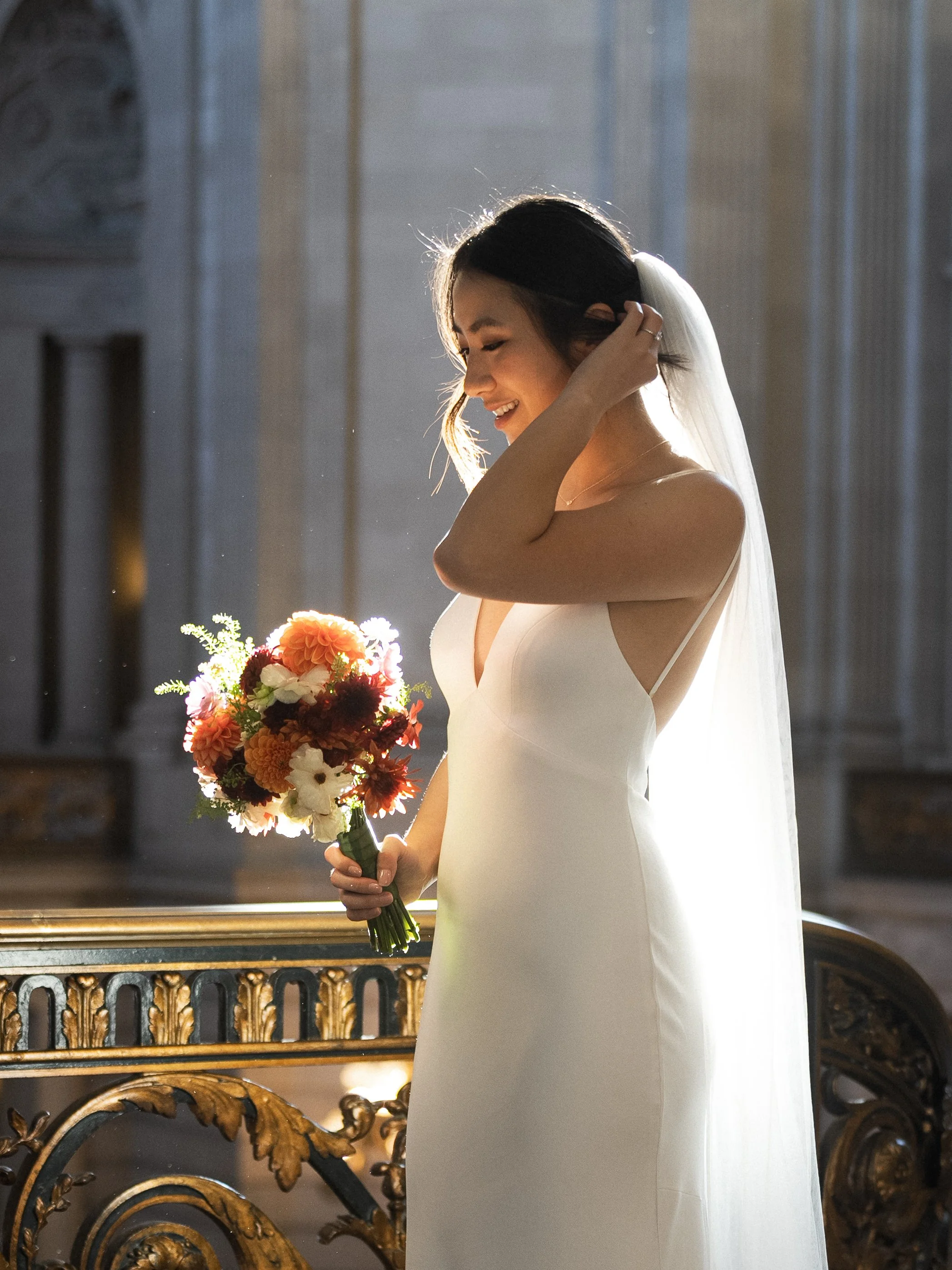 A bride in a white wedding dress holding a colorful bouquet of flowers and smiling inside a grand, ornate building.