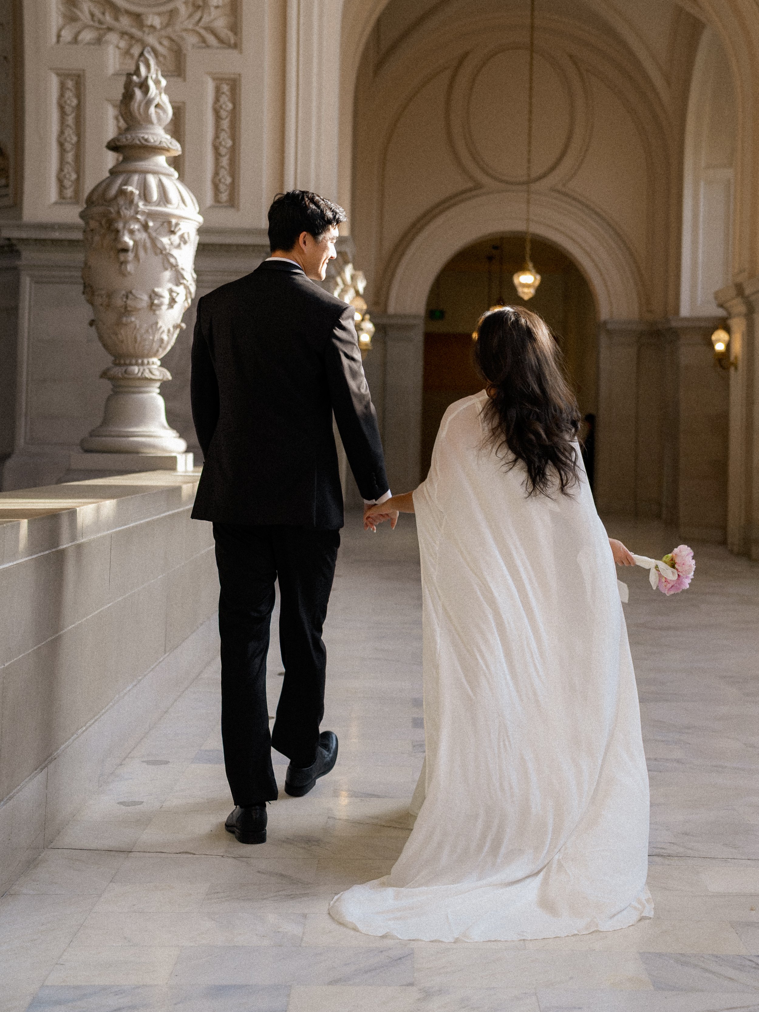 A bride in a white wedding dress holding a pink flower, walking with a groom in a black suit inside a grand, ornate hall with tall arches and decorative columns.