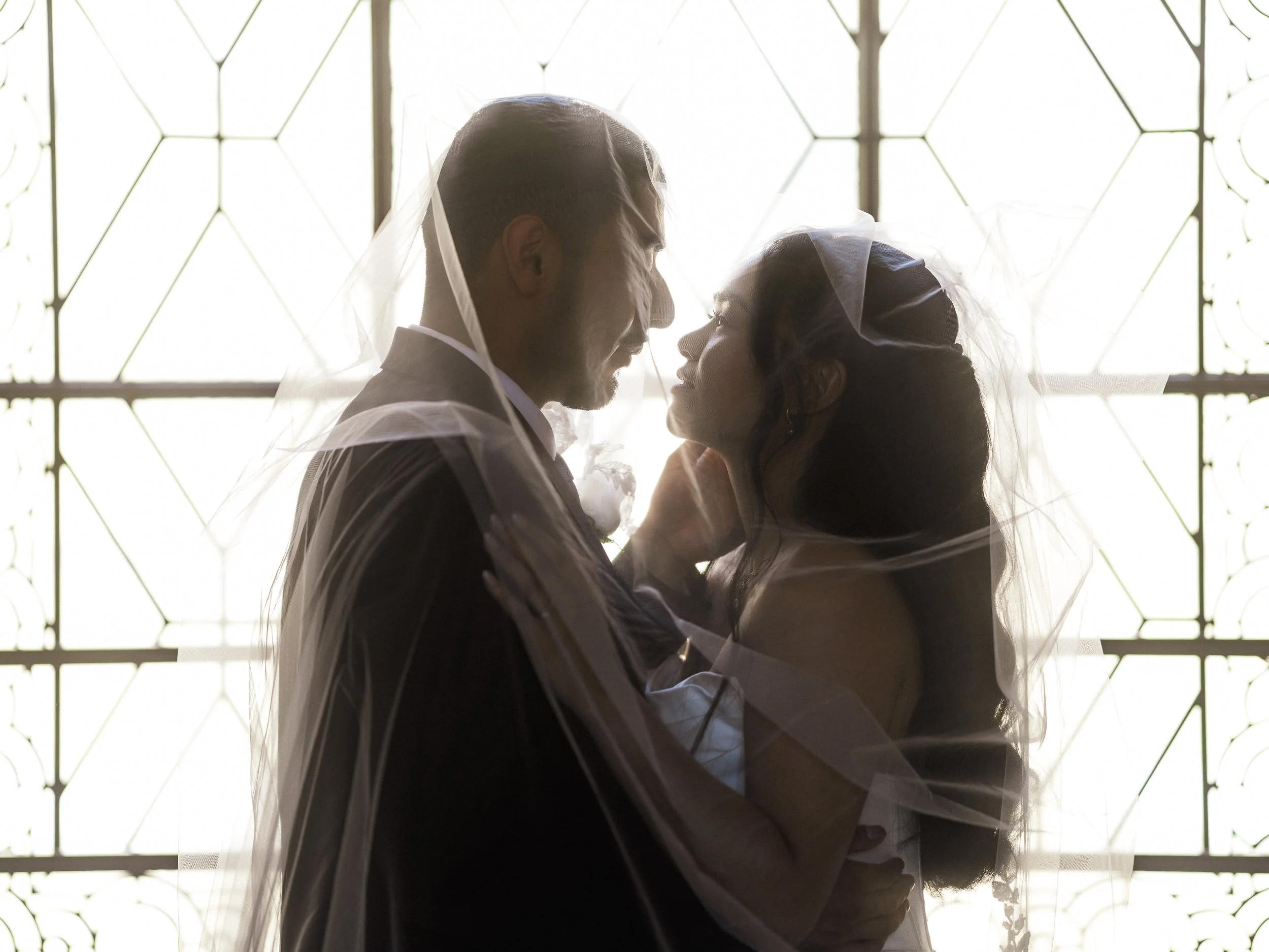 A bride and groom kissing behind a veil in front of a stained glass window.