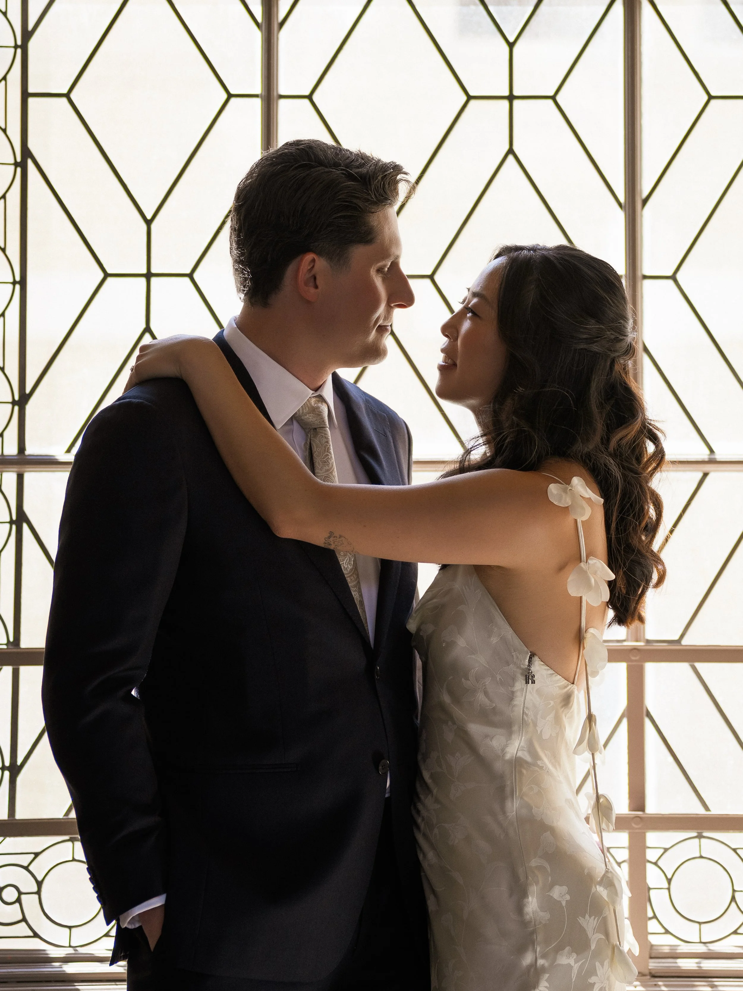 A couple dressed in formal attire, standing close together and looking into each other's eyes indoors with decorative patterned windows in the background.