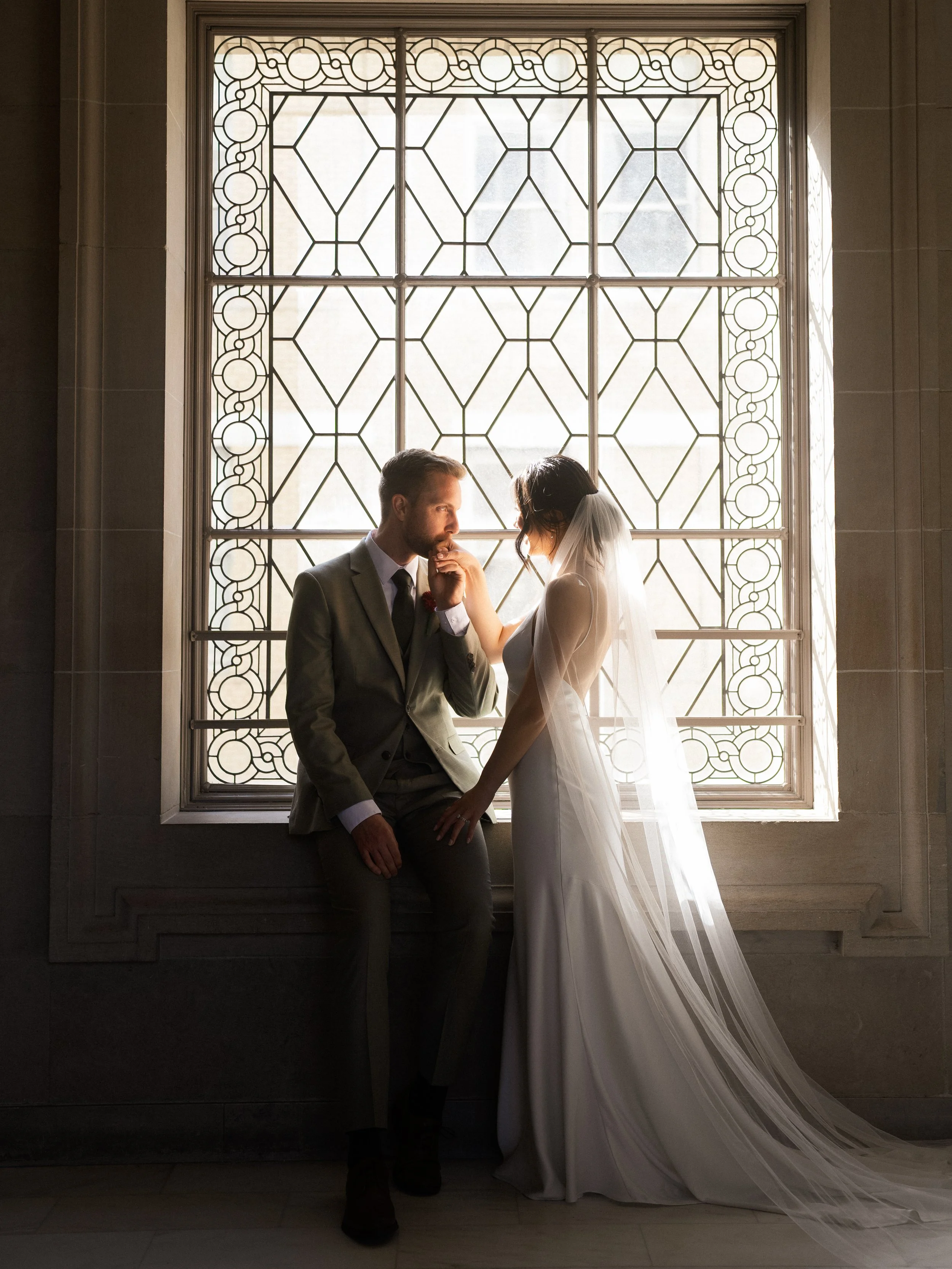A bride and groom in wedding attire in front of a decorative window, with the bride touching the groom's face as they gaze at each other.
