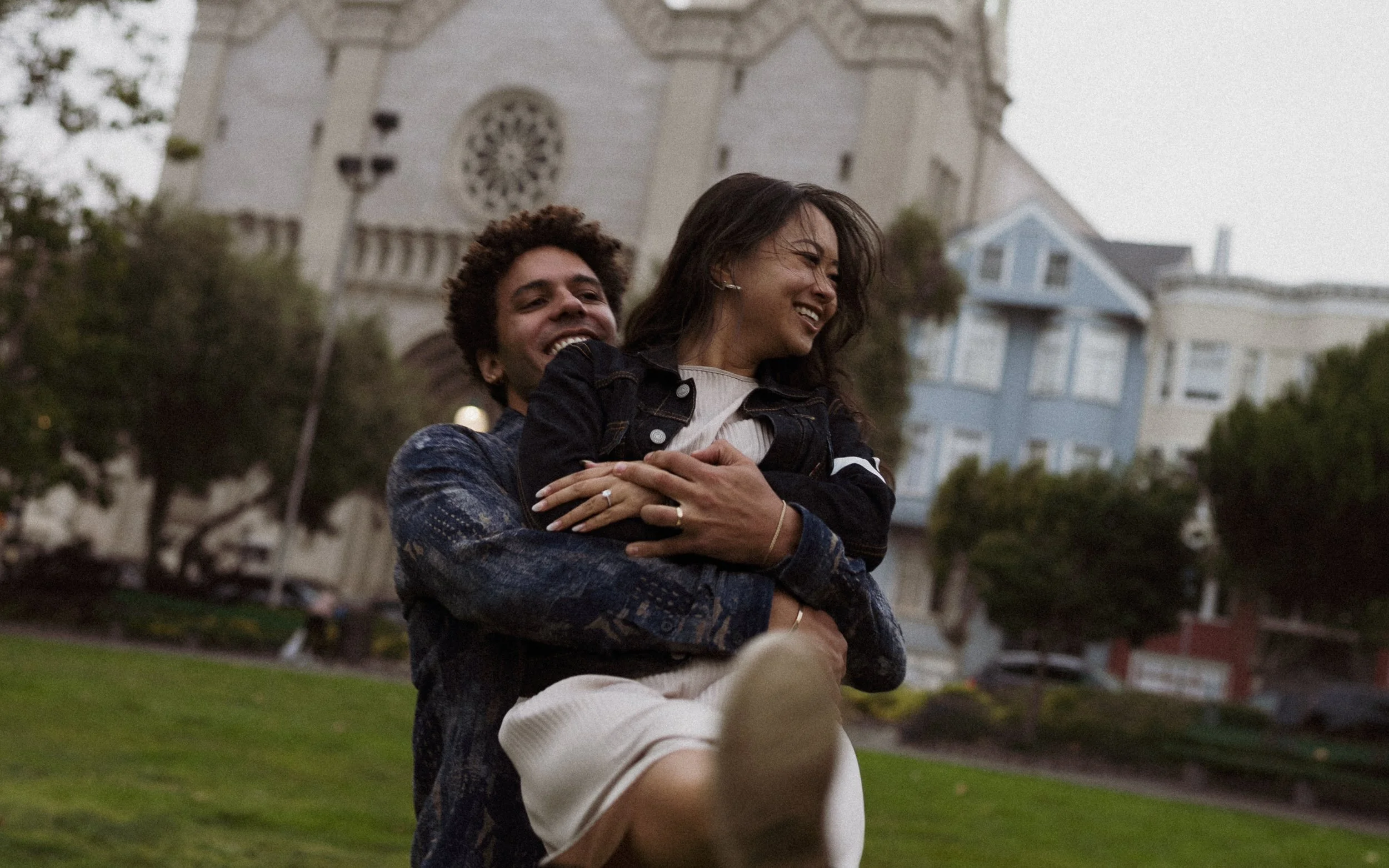 A couple is outdoors, with the man carrying the woman on his back, both smiling and enjoying themselves. In the background, there's a church with a large rose window and residential houses, and trees around them.