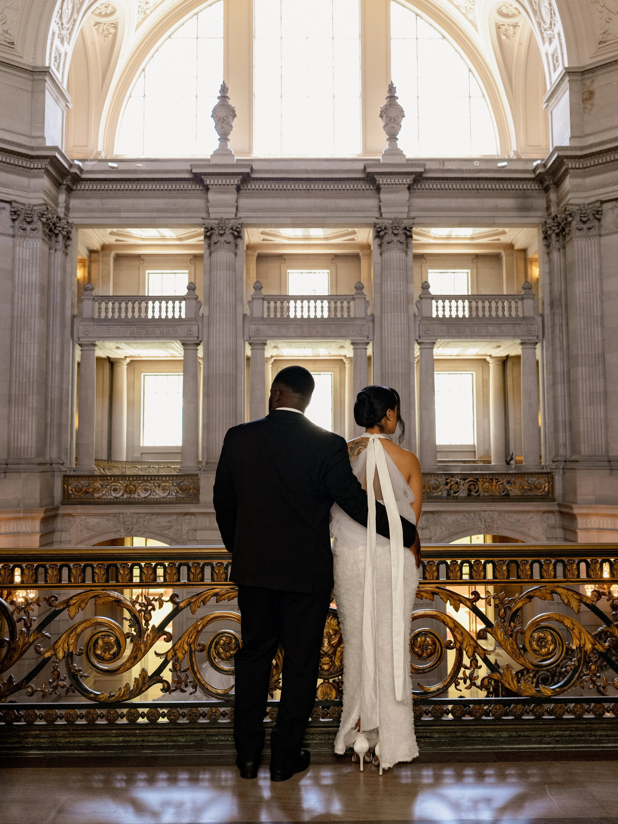A couple dressed in wedding attire standing on a balcony inside a grand, historic building with ornate architecture, large windows, and classical columns.