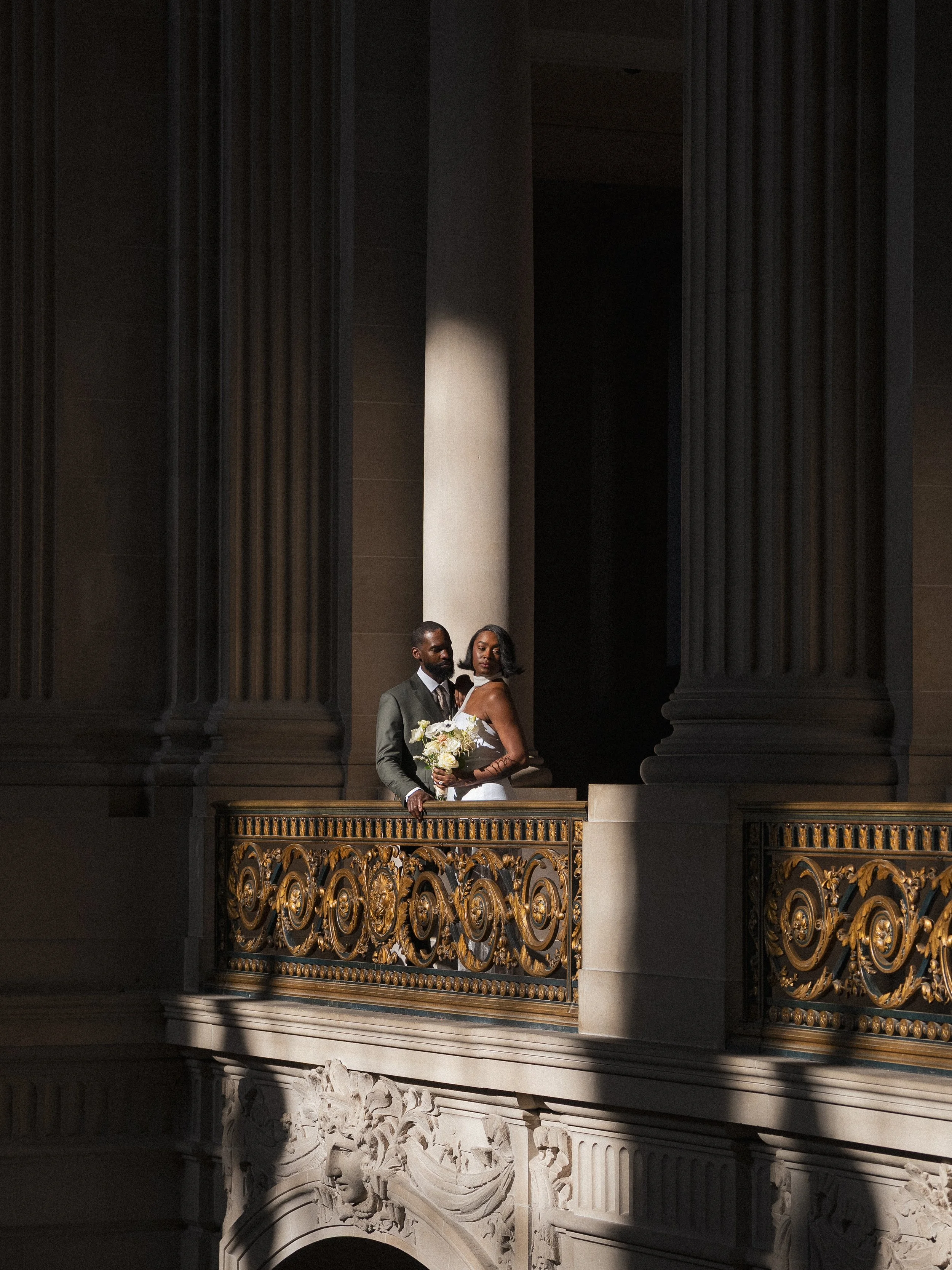 A couple dressed in wedding attire standing on a balcony inside a grand, elegant building with large columns and ornate golden railing.