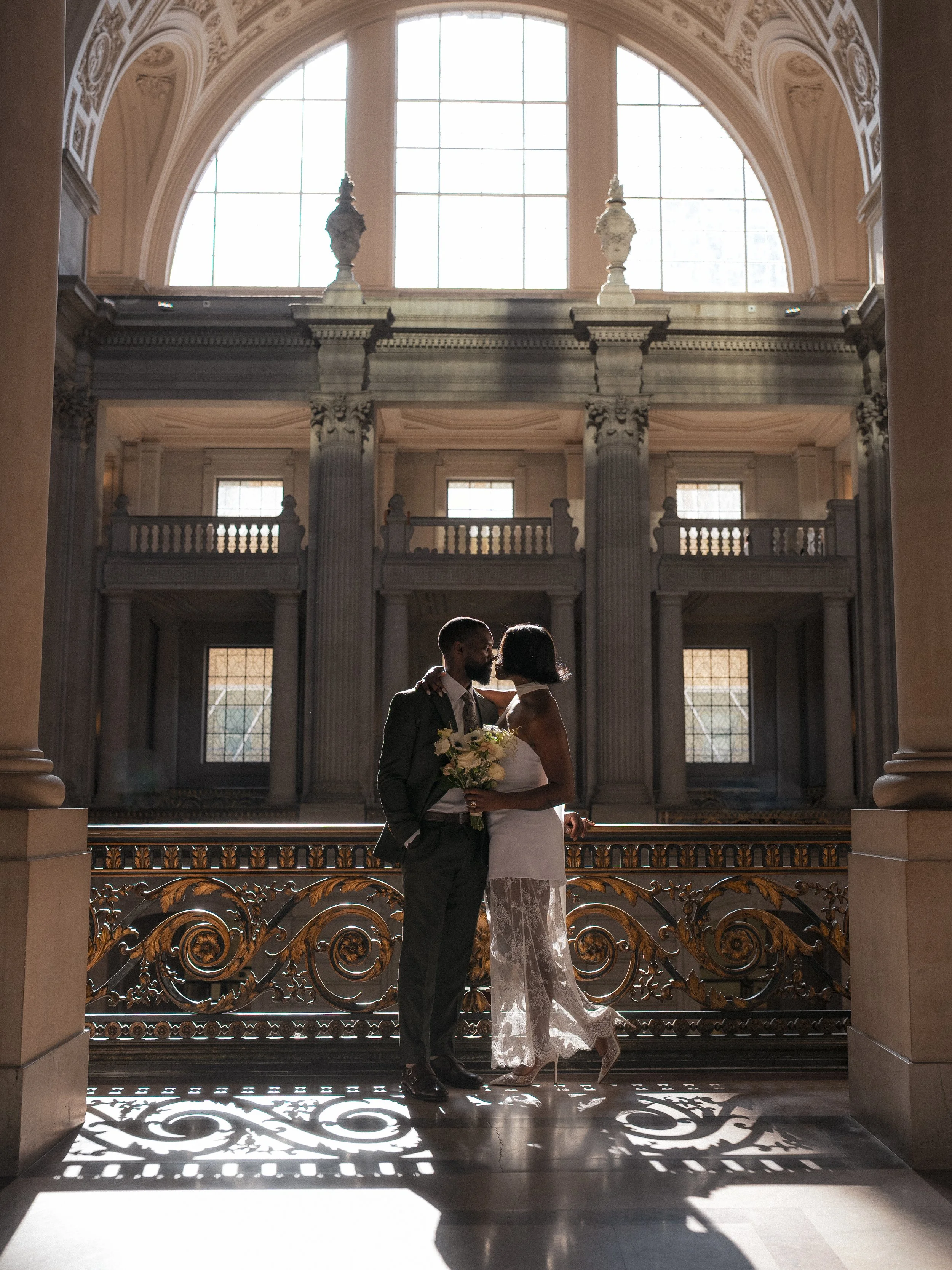 A couple embracing in wedding attire, standing on a grand staircase inside an ornate, historic building with large windows and intricate architectural details, backlit by sunlight.