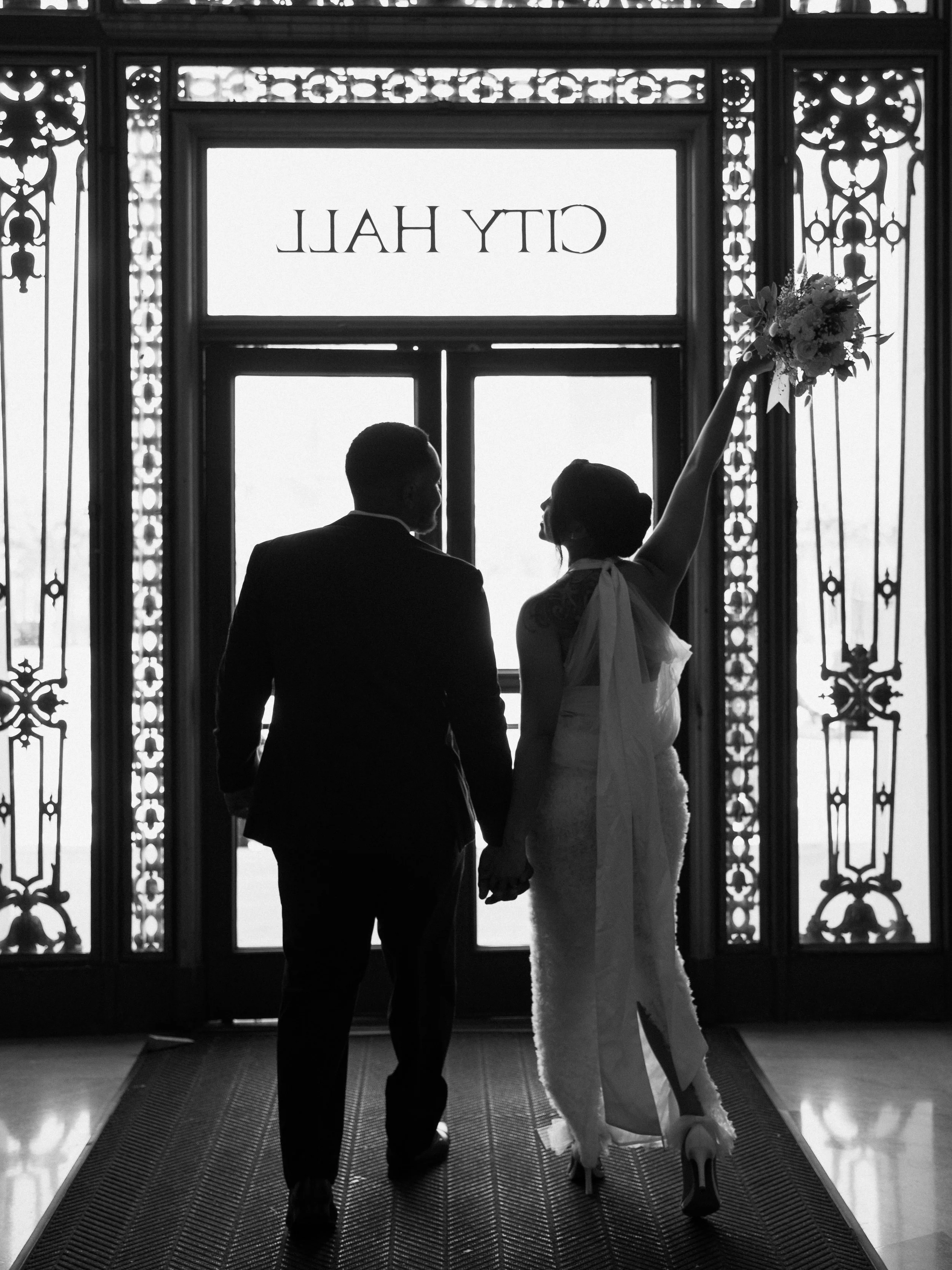 A black and white photo of a bride and groom holding hands and facing a window at CIT YHALL. The bride is holding a bouquet in her right hand and wearing a strapless dress with high heels. The groom is dressed in a suit.
