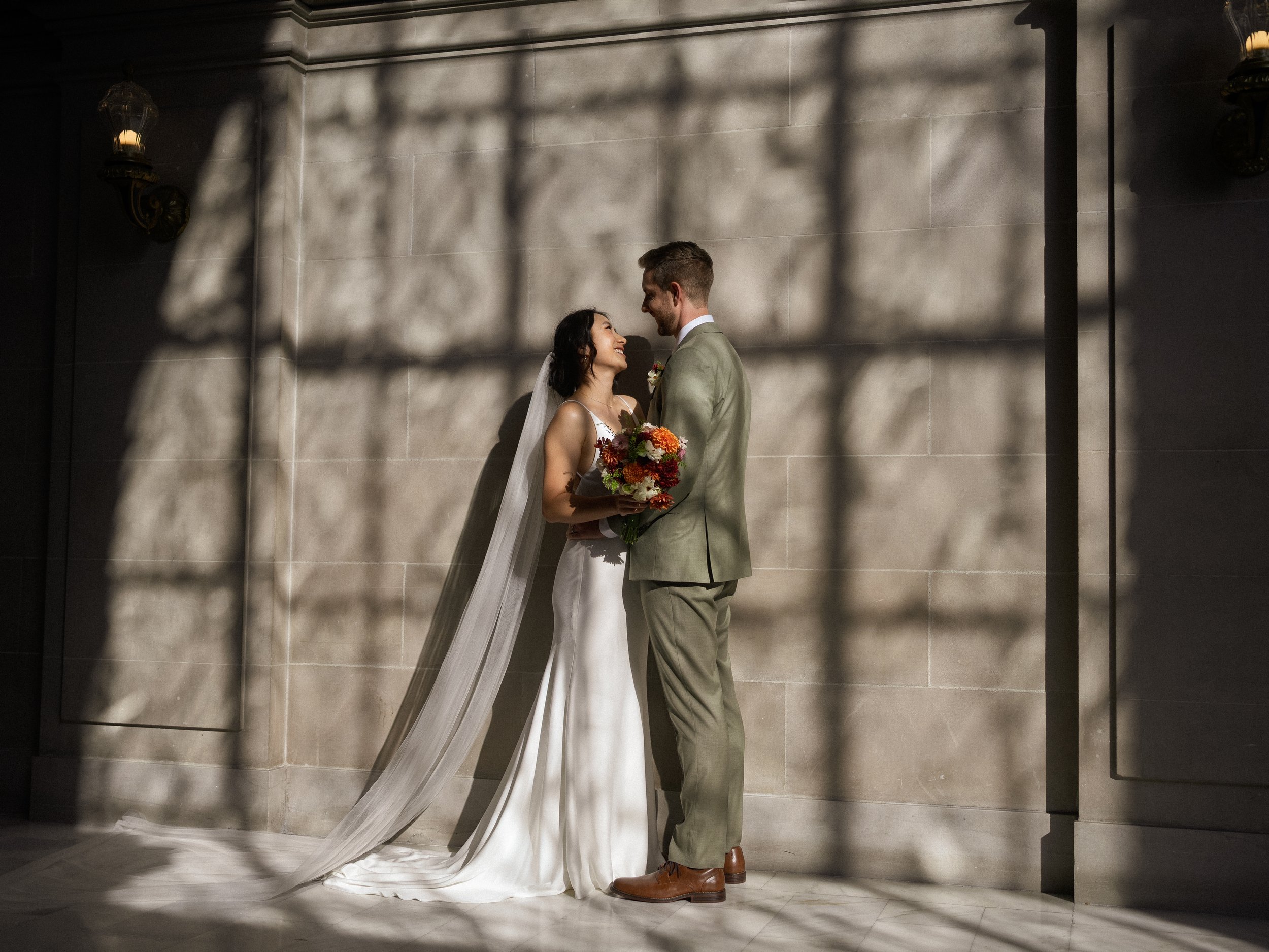 A bride and groom standing close together indoors, holding a bouquet of flowers, with shadows of branches cast on the wall behind them.