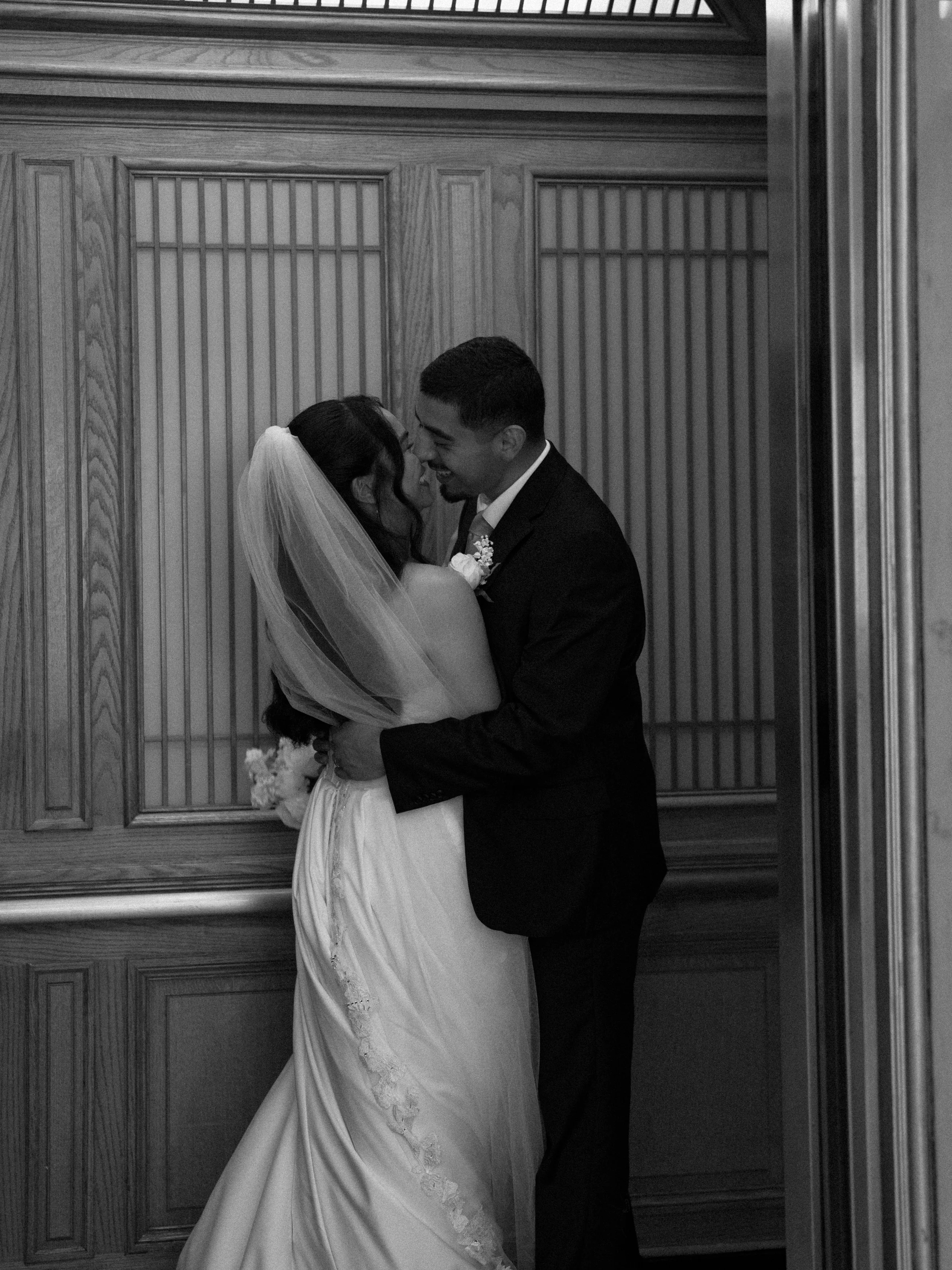A bride and groom are embracing and smiling at each other in a wood-paneled room, with the bride wearing a wedding dress and veil, and the groom in a tuxedo.